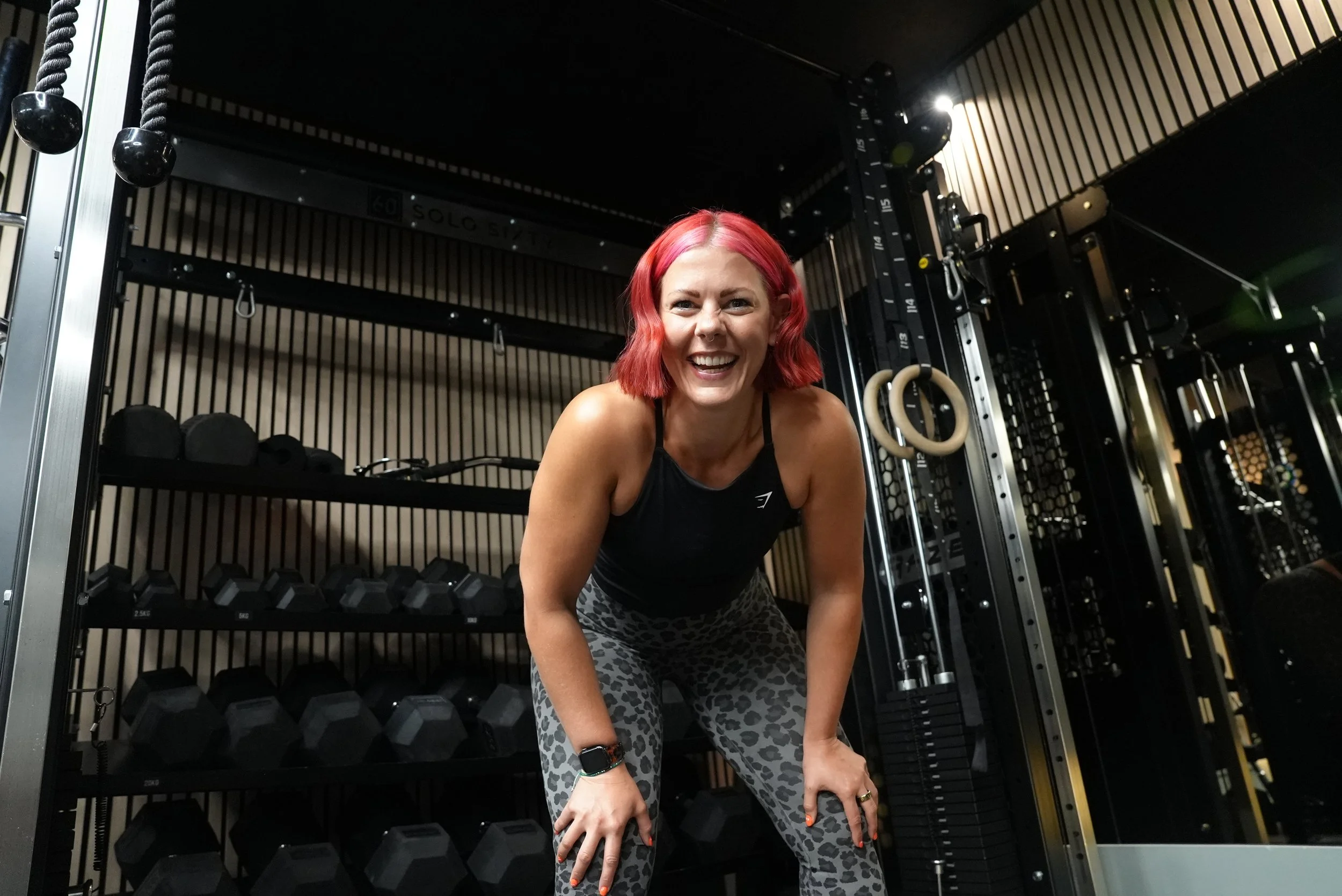 Woman with red hair smiling in a fitness gym, surrounded by weights and exercise equipment.