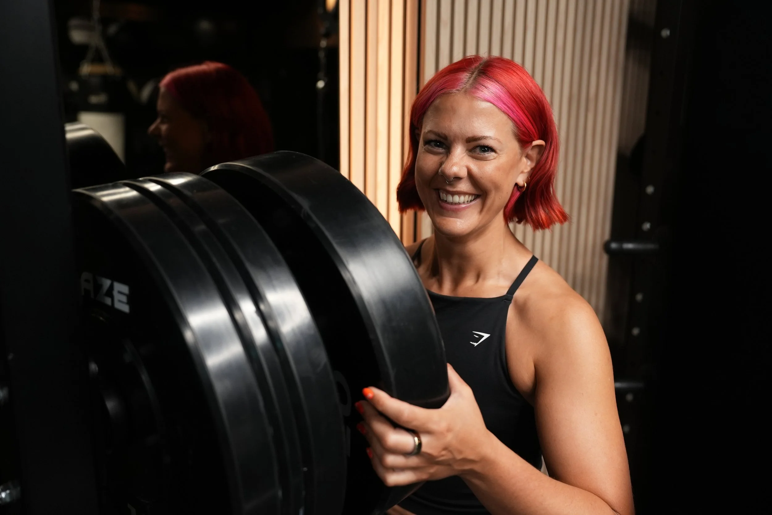 A woman with pink hair in a black tank top smiling in a gym, holding a large weight plate.