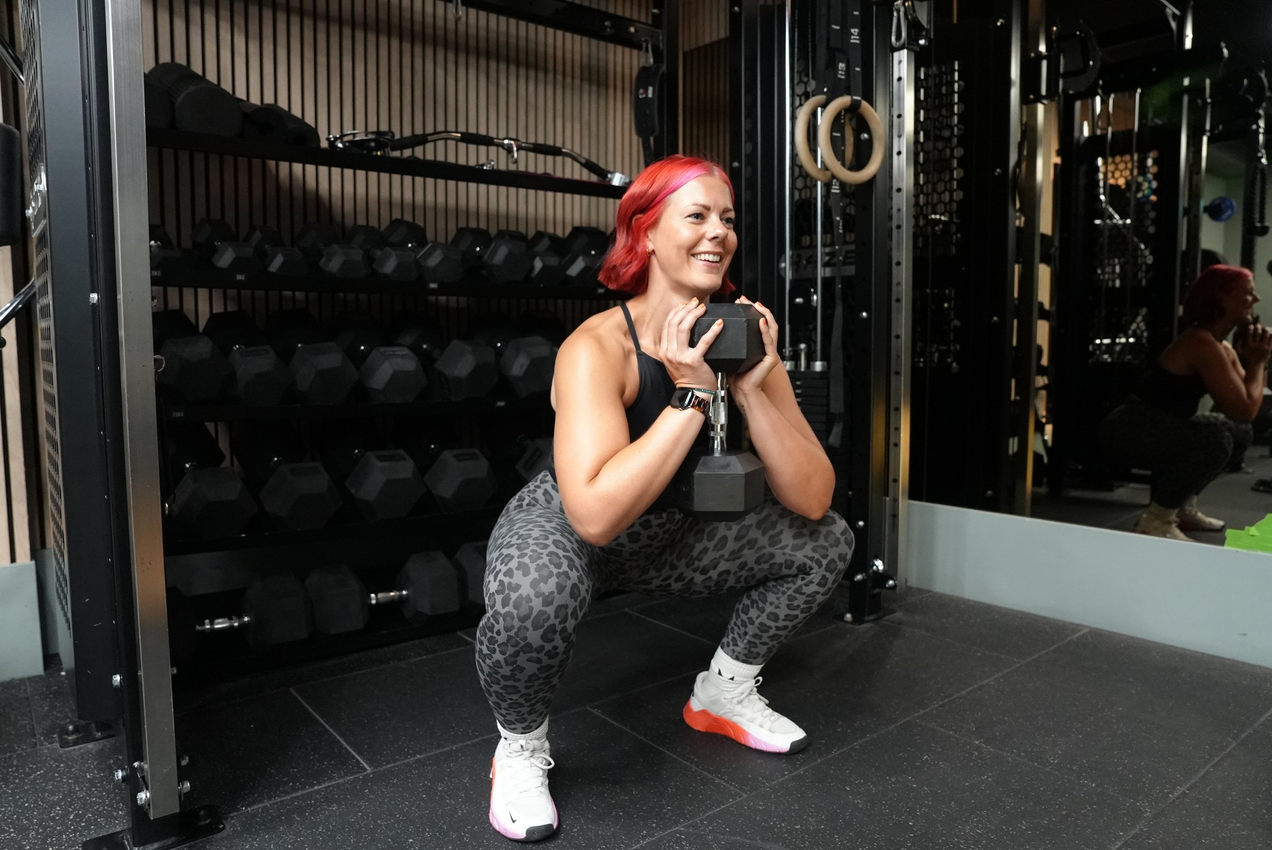 A woman with red hair doing squats in a gym, holding a dumbbell close to her chest, surrounded by workout equipment including free weights and gymnastic rings.