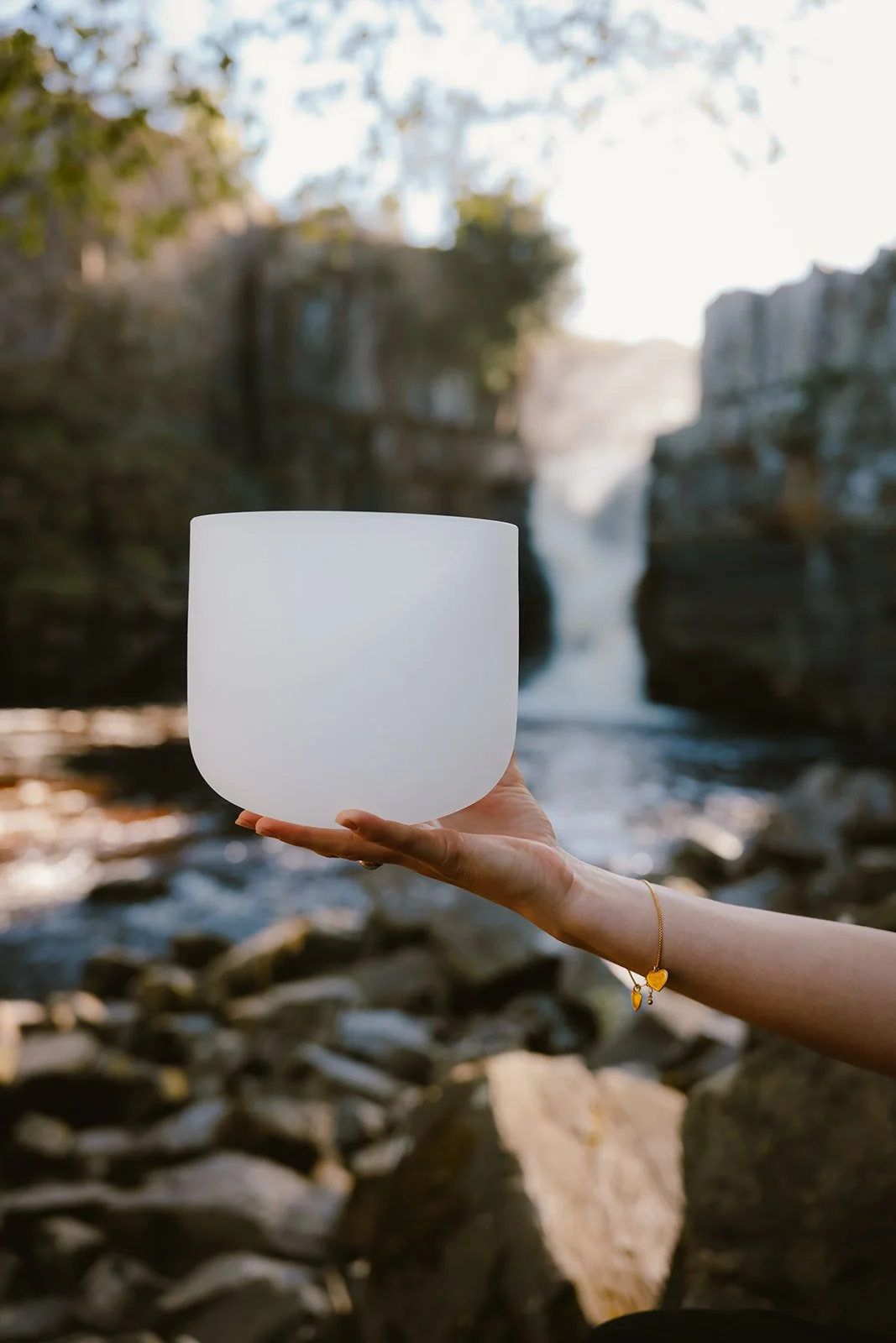 A person holding a glowing white bowl in front of a waterfall and rocky river with trees and a cloudy sky in the background.