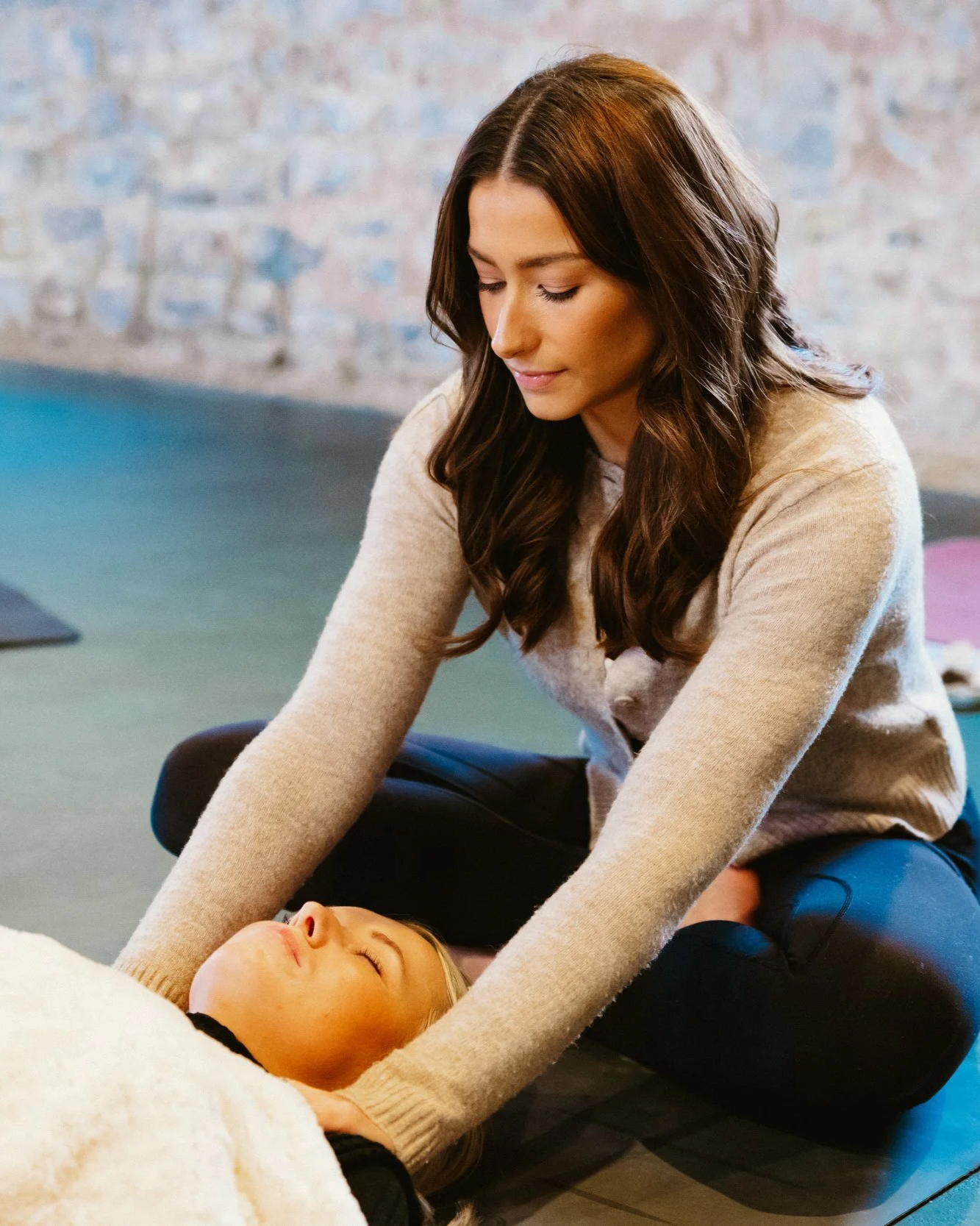 A woman in a beige sweater performs a gentle touch breast massage on a lying woman with blonde hair, in a calming indoor setting.