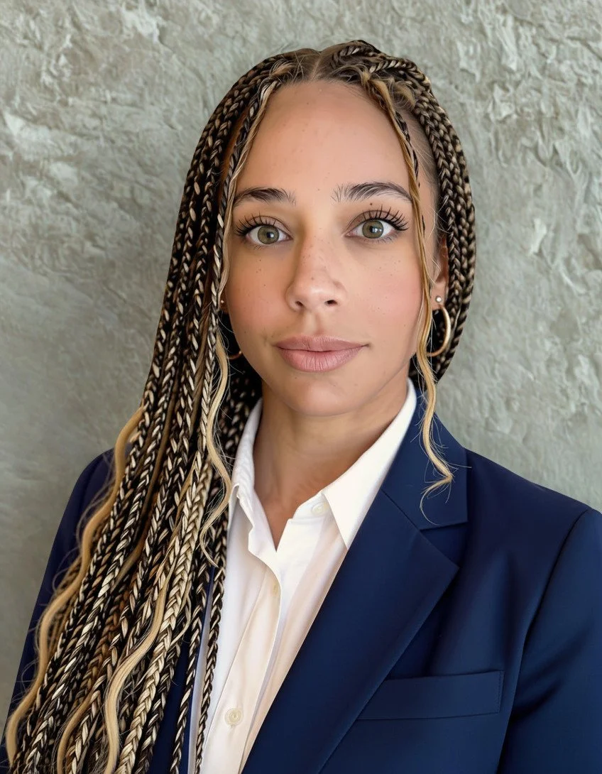 A woman with long braided hair wearing a navy blazer and white shirt, standing in front of a textured light-colored wall.