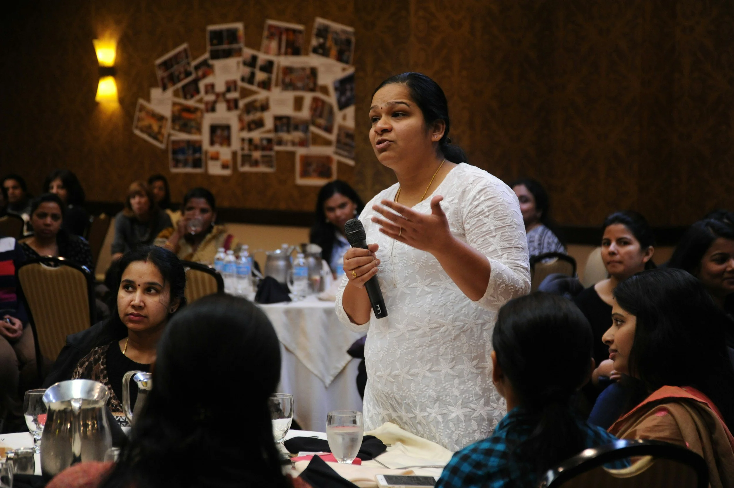 A woman in a white dress holding a microphone and speaking to a group of people at a conference or seminar.