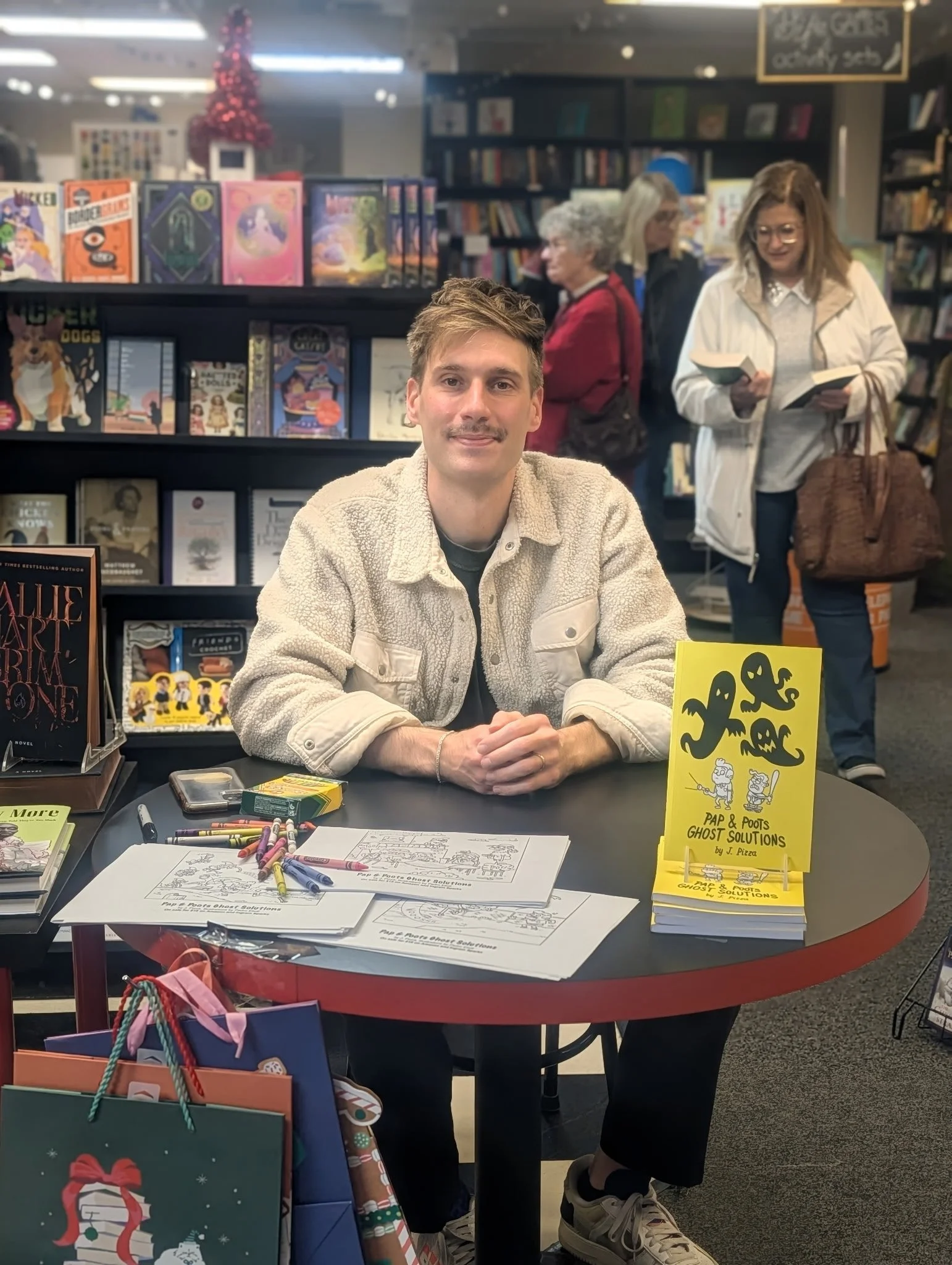 A man with a mustache and light brown hair, wearing a beige fleece jacket, sitting at a table with coloring books, pages, markers, and a yellow book titled 'PAP & POOFS GHOST SOLUTIONS' by J. Pizza. The background shows bookshelves, several women browsing, and a Christmas tree.