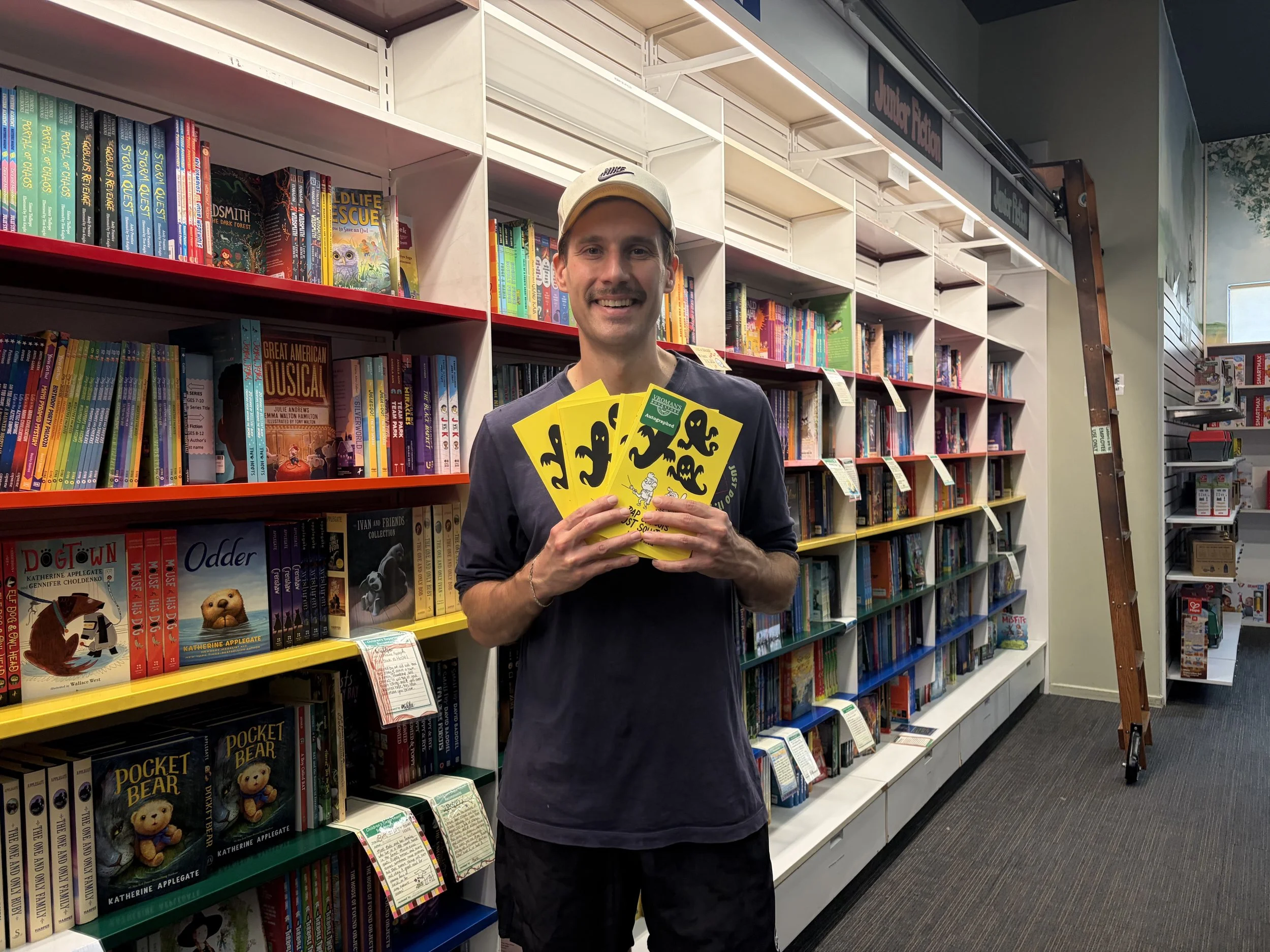 A man wearing a beige Nike cap and black shirt is smiling and holding several yellow posters with black octopus illustrations in a bookstore aisle. The background shows tall bookshelves filled with colorful children's books.