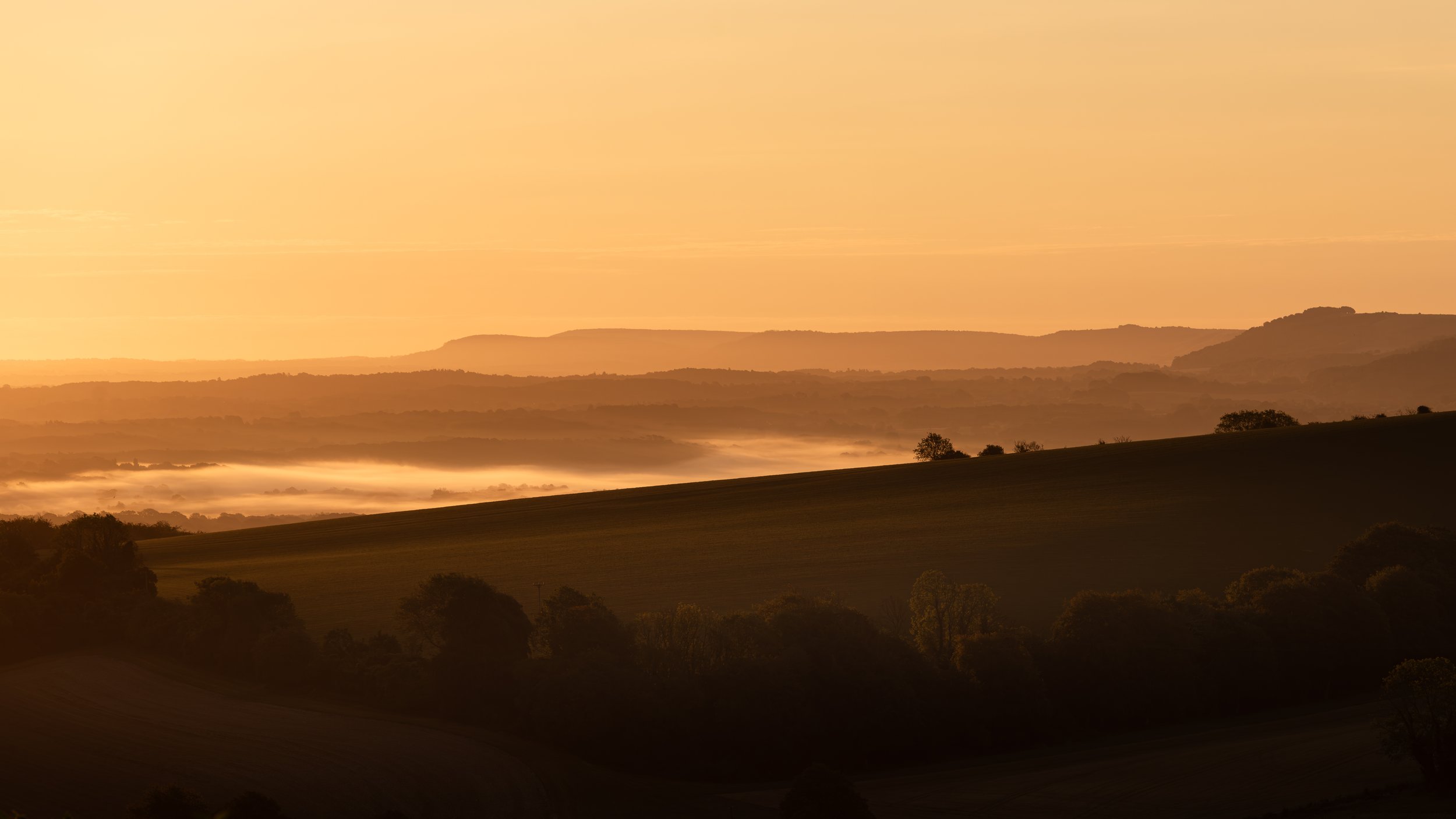 A landscape scene at sunrise or sunset with rolling hills, trees, and a sky with a warm, orange glow.