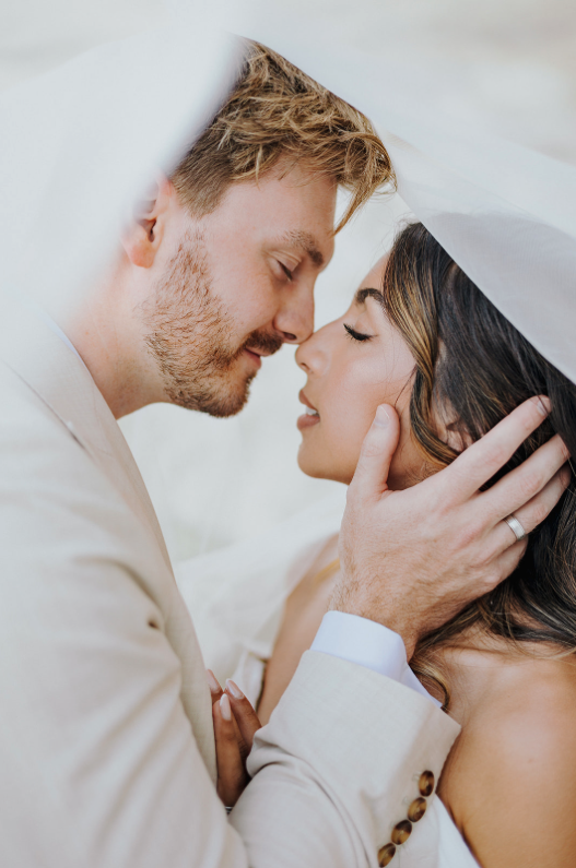 A romantic moment between a man and a woman, with their foreheads touching and eyes closed, under a white cloth or veil, conveying intimacy and love.