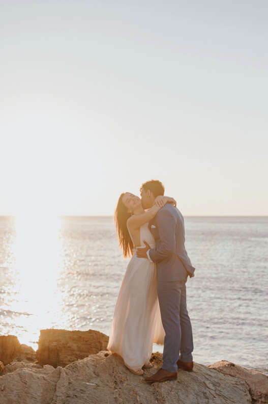 A couple dressed in wedding attire embracing on a rocky shoreline during sunset.
