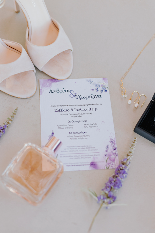 Wedding invitation surrounded by white shoes, pearl earrings and necklace, perfume bottle, and purple lavender flowers.