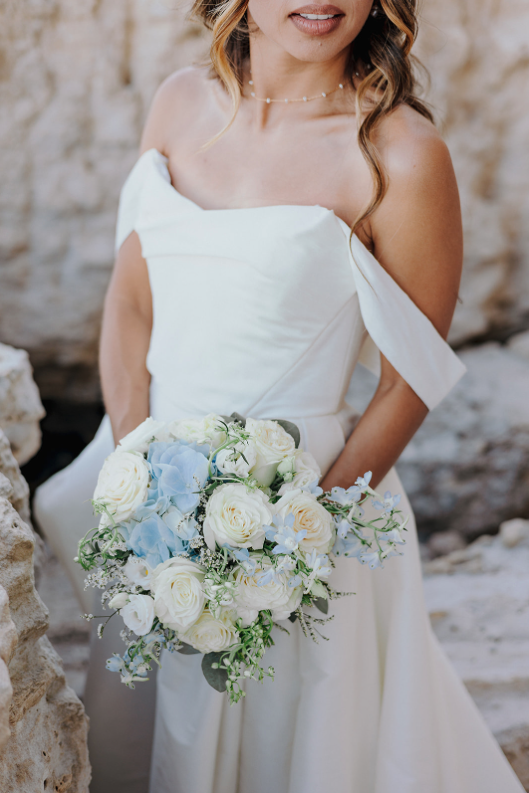 Woman in a white off-shoulder wedding dress holding a bouquet of white and pale blue flowers, standing outdoors with rocks in the background.