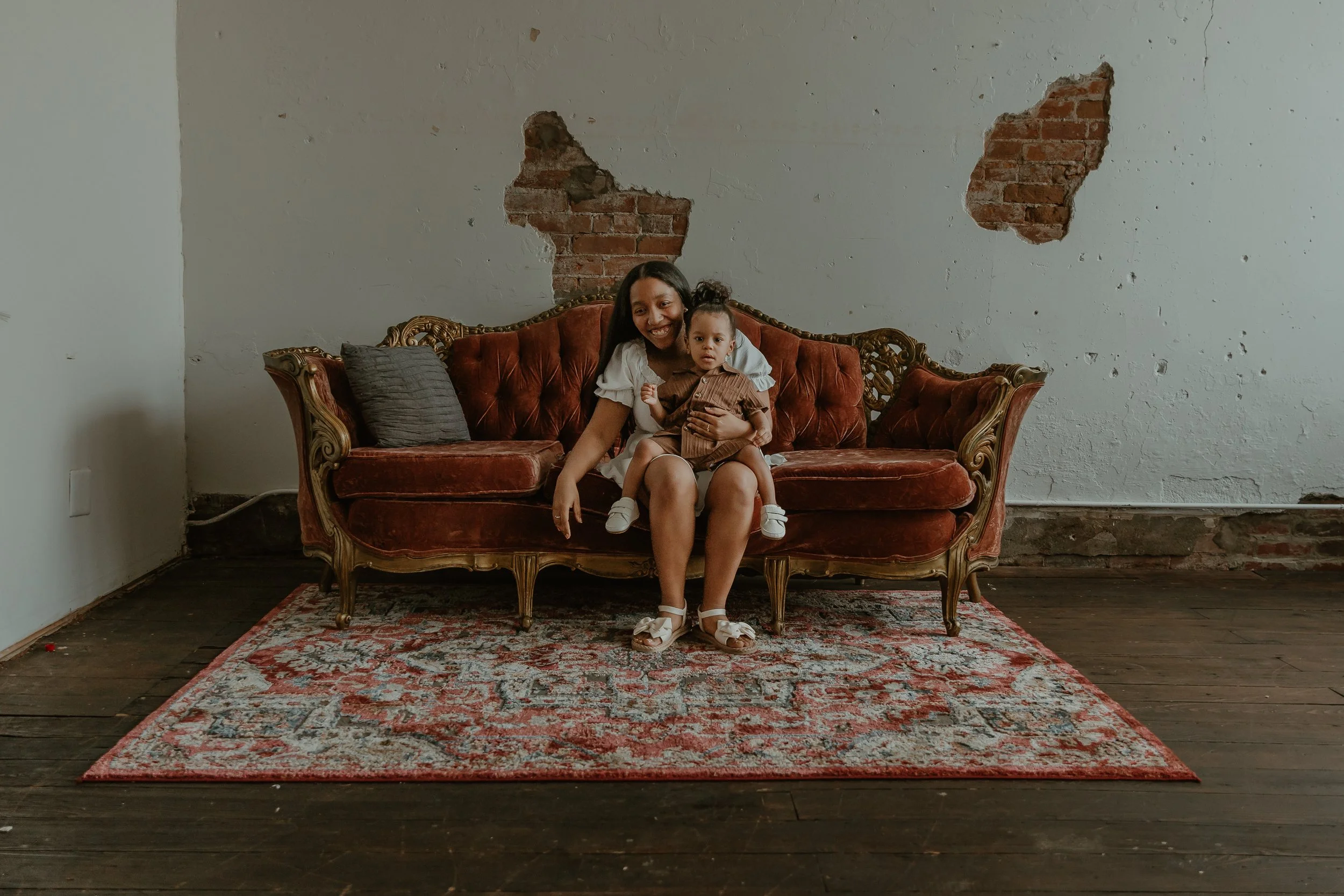 A woman and a child sitting on an ornate vintage sofa in a room with a distressed and partially exposed brick and plaster wall behind them. The woman has long dark hair and is smiling, while the child has curly hair and is sitting on her lap looking at the camera.