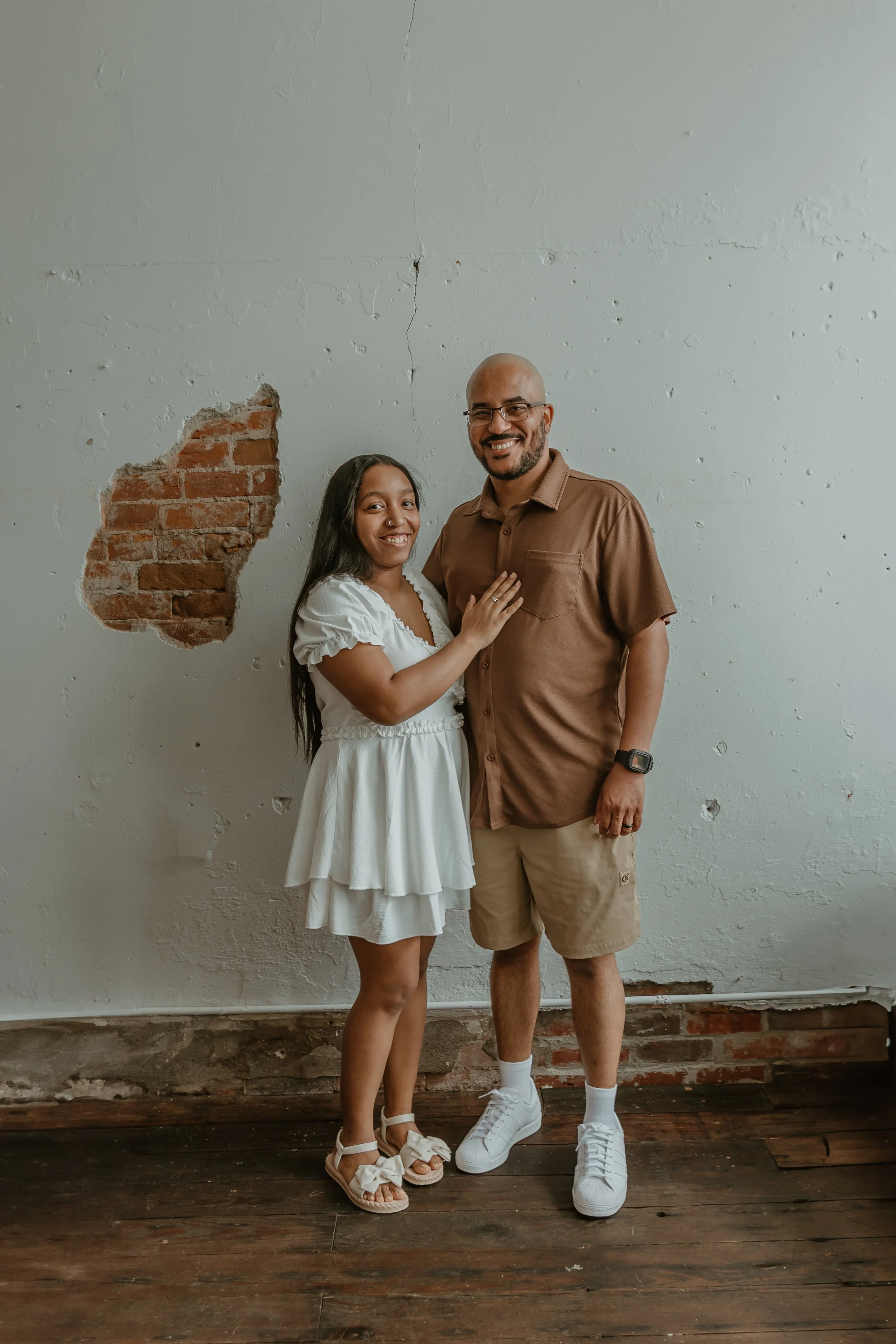 A smiling woman in a white dress and a man in a brown shirt and beige shorts stand together indoors against a white wall with a brick section. They are smiling at the camera.