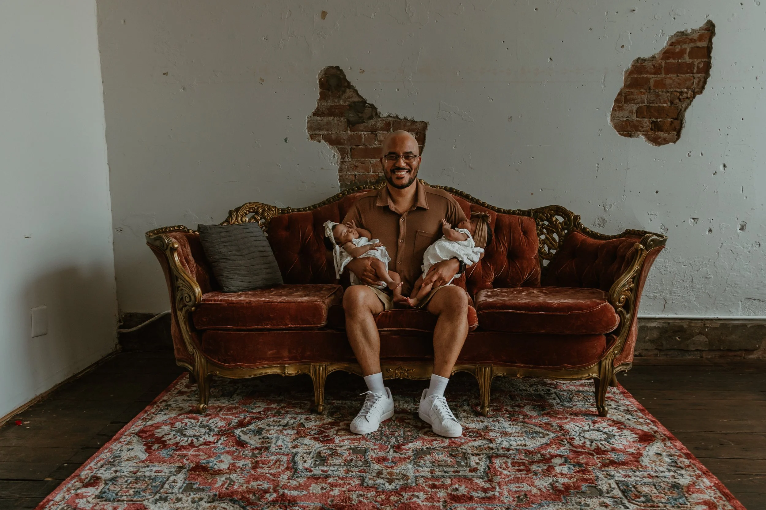 A man with glasses and a beard sitting on an ornate vintage red sofa, smiling, holding two sleeping babies dressed in white, in a room with a distressed white wall with exposed brick patches, a red patterned rug, and wooden floor.