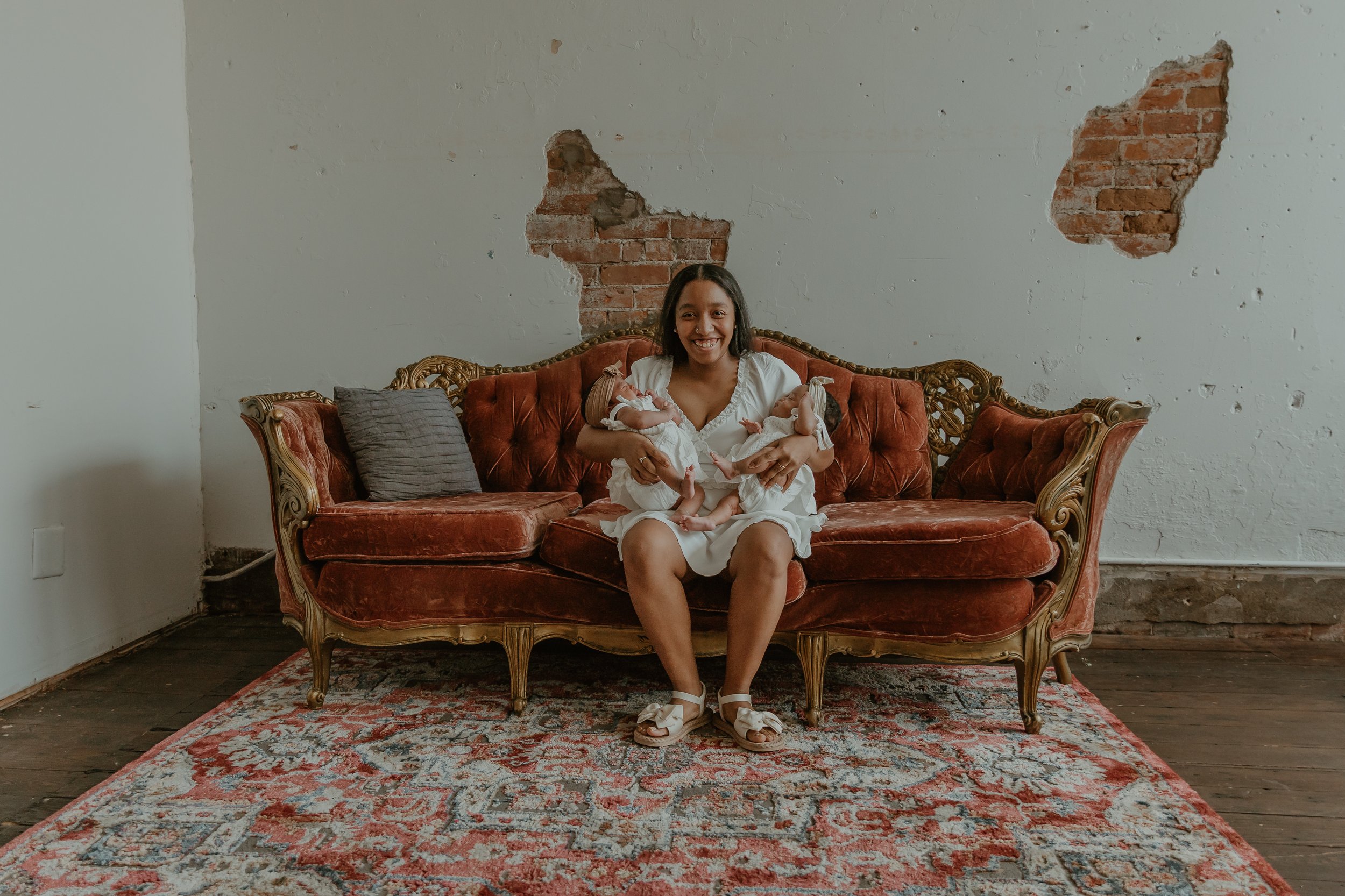 A woman sitting on an ornate red velvet vintage sofa, holding and smiling at two newborn babies in white clothing. The sofa is placed on a patterned rug on a wooden floor against a wall with exposed brick patches and worn paint.