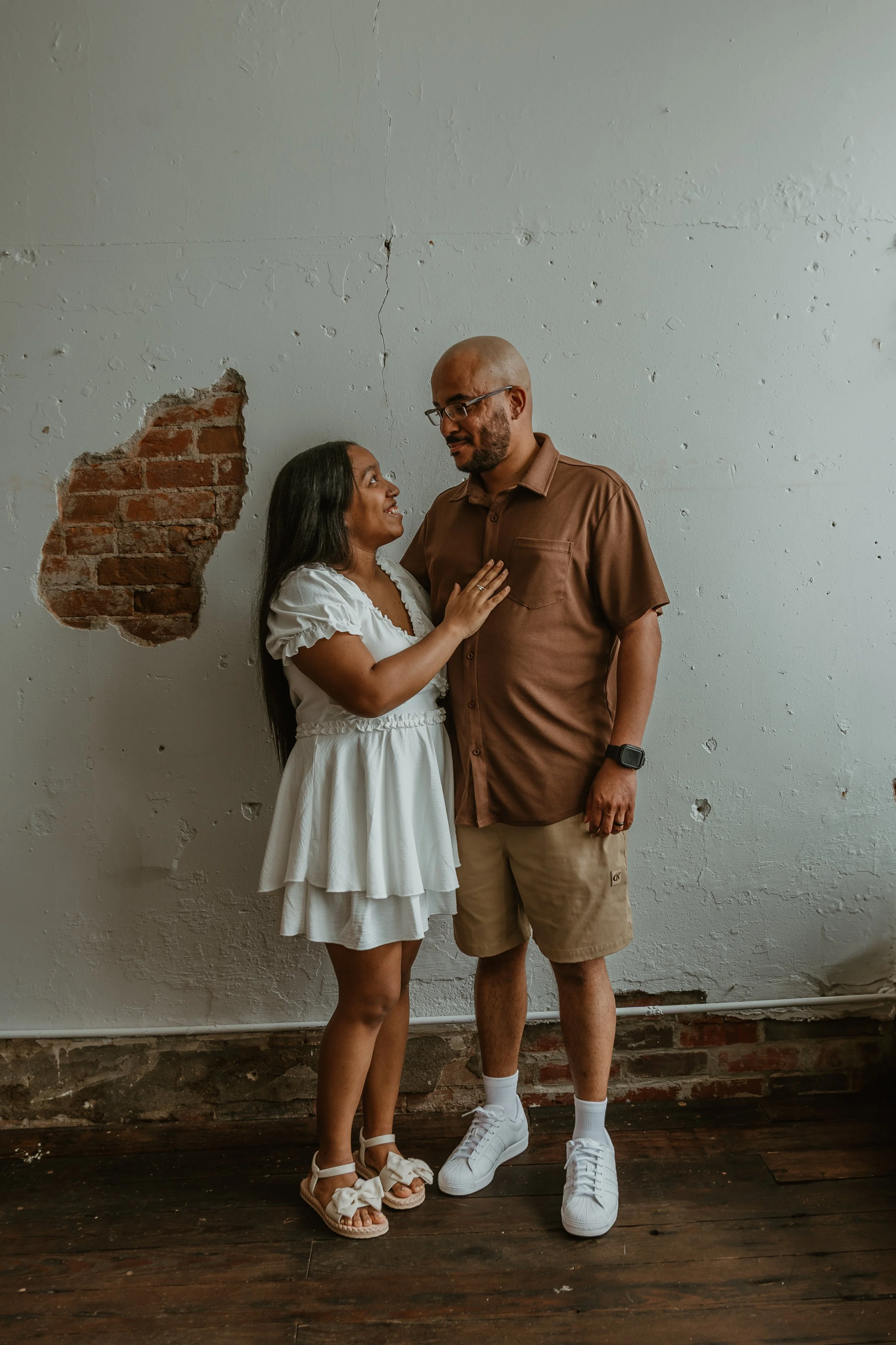 A man and woman standing close together in front of a distressed white wall with exposed brick. The woman is wearing a white dress and sandals, while the man is dressed in a brown shirt, beige shorts, and white sneakers. They are looking at each other and smiling.