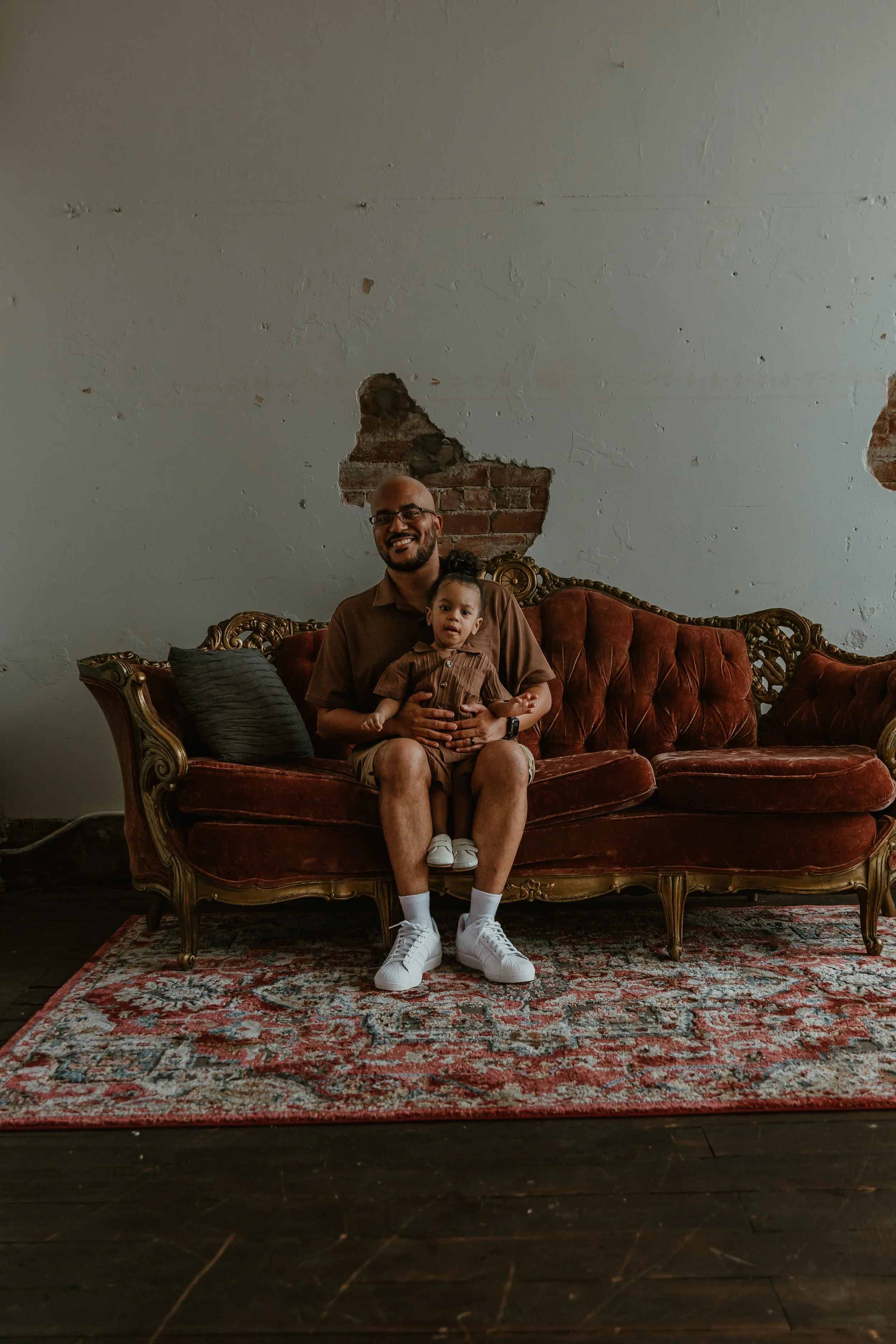 A man and a young child sit on an ornate vintage sofa in a room with a distressed wall, a patterned rug, and wooden flooring.