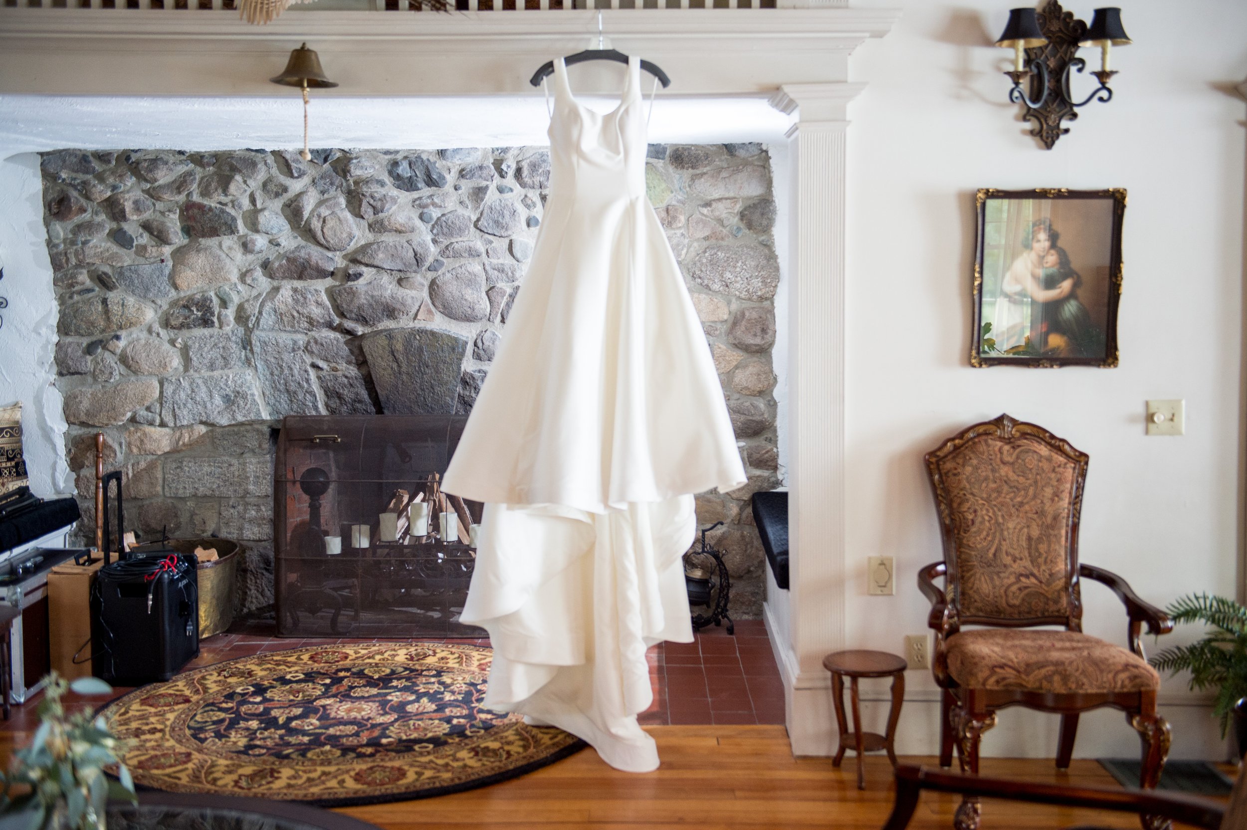 A white wedding dress hanging from a black hanger in front of a stone fireplace inside a living room, with classic wall sconces, a framed painting on the wall, an ornate armchair, a patterned rug, and various household items.