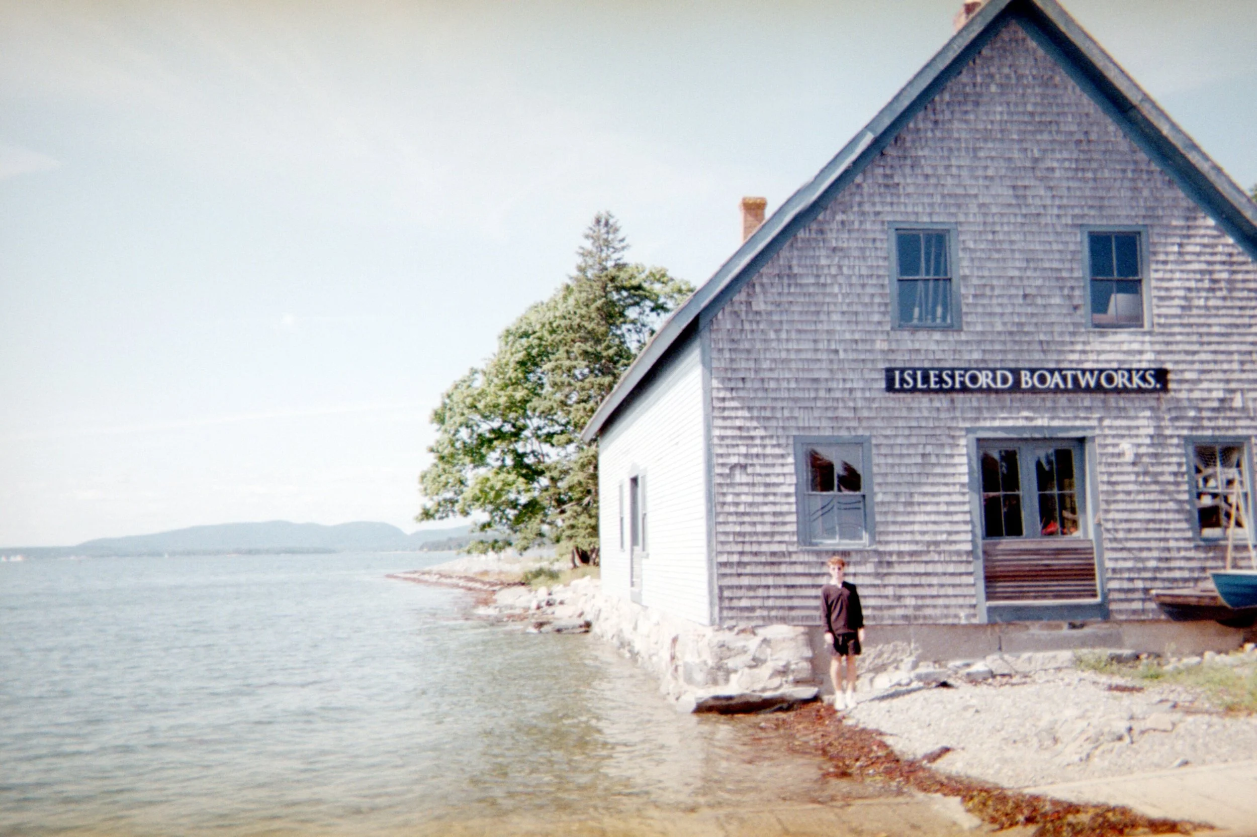 A wooden house with a sign that reads "Islesford Boatworks" on the side. The house is next to a body of water with a rocky shoreline and a person standing in front of it. There are some trees and distant hills visible in the background.