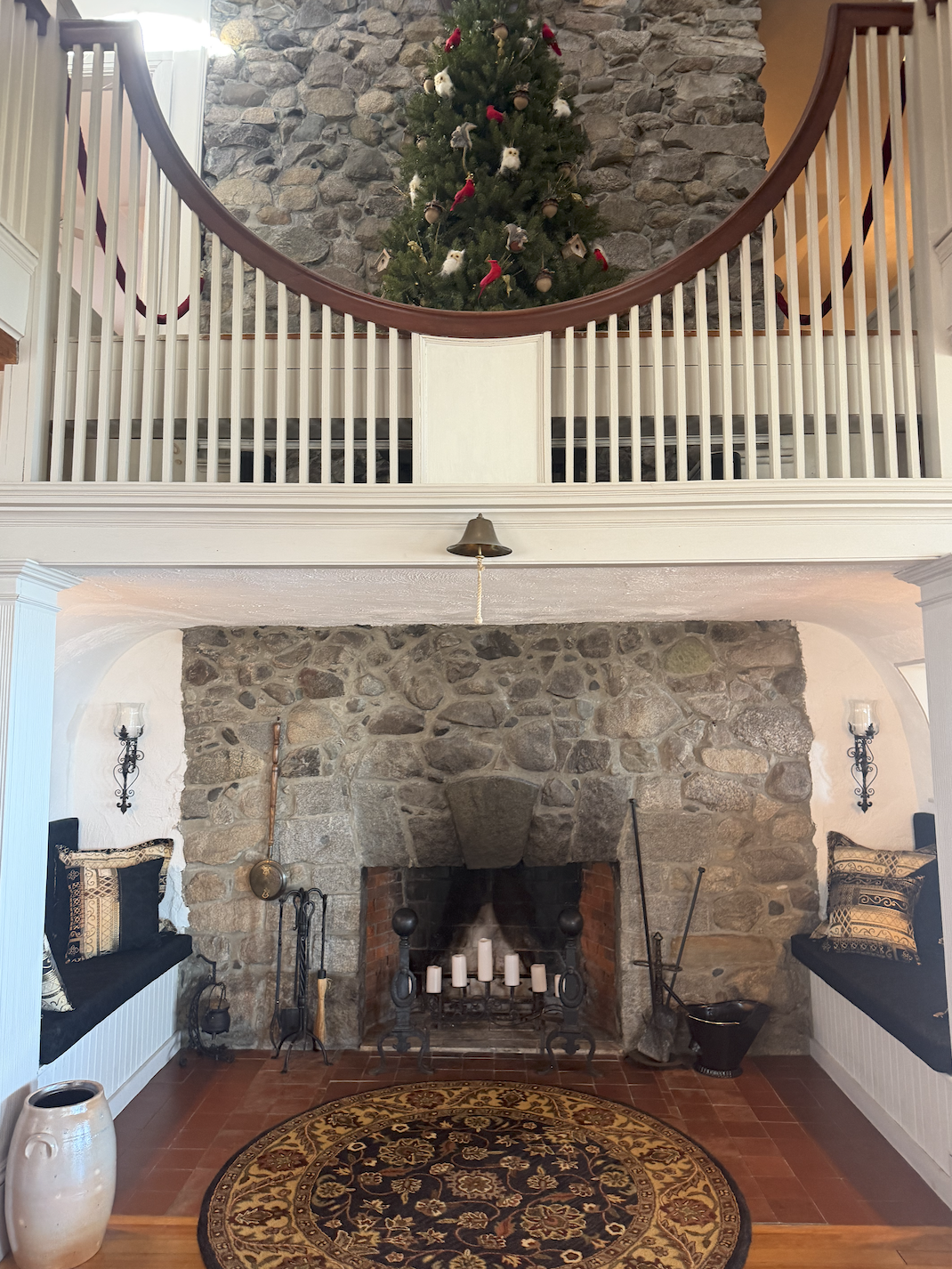 Living room with stone fireplace, decorated for Christmas with greenery on a second-floor balcony, a round rug, and built-in window seats with pillows.