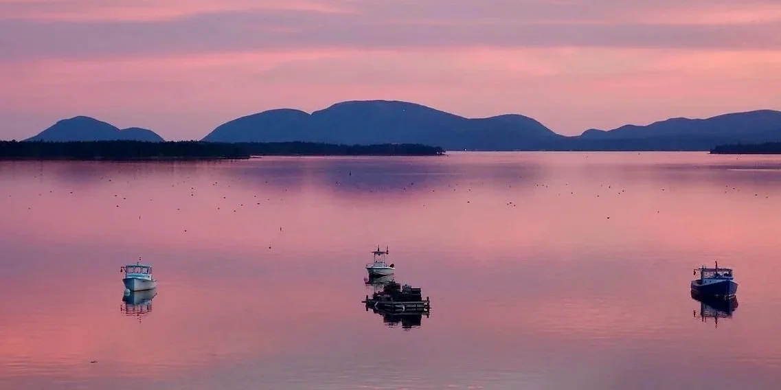 Frenchman's Bay, Bar Harbor, and Acadia National Park at sunset with four boats floating, pink and purple sky, distant mountains in the background.