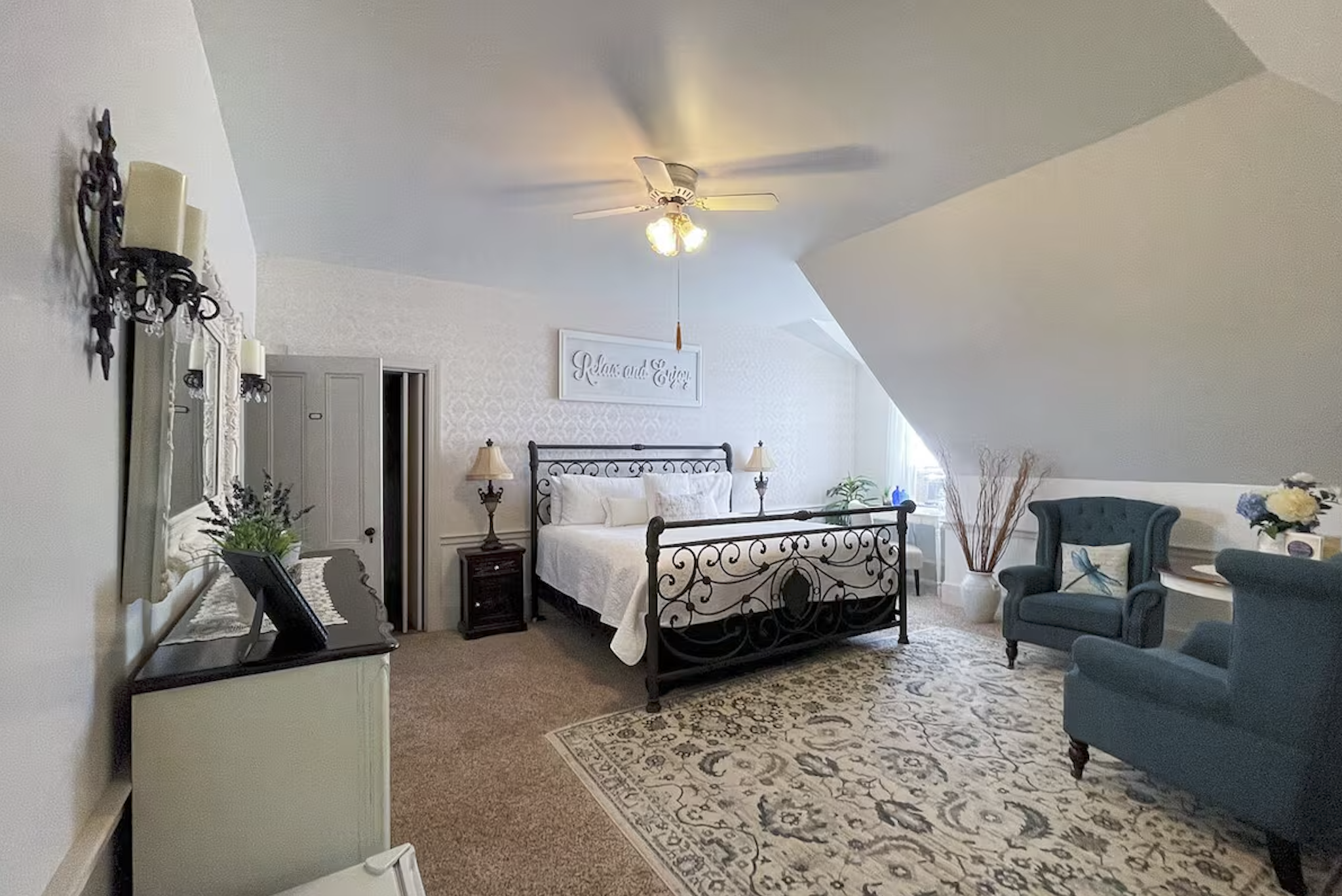 A cozy bedroom with a white ceiling fan, a bed with a wrought iron frame, and vintage-style furnishings including a dresser, lamps, and a seating area with a blue chair. Decor includes a framed sign that reads 'Relax and Enjoy,' plants, and floral arrangements.