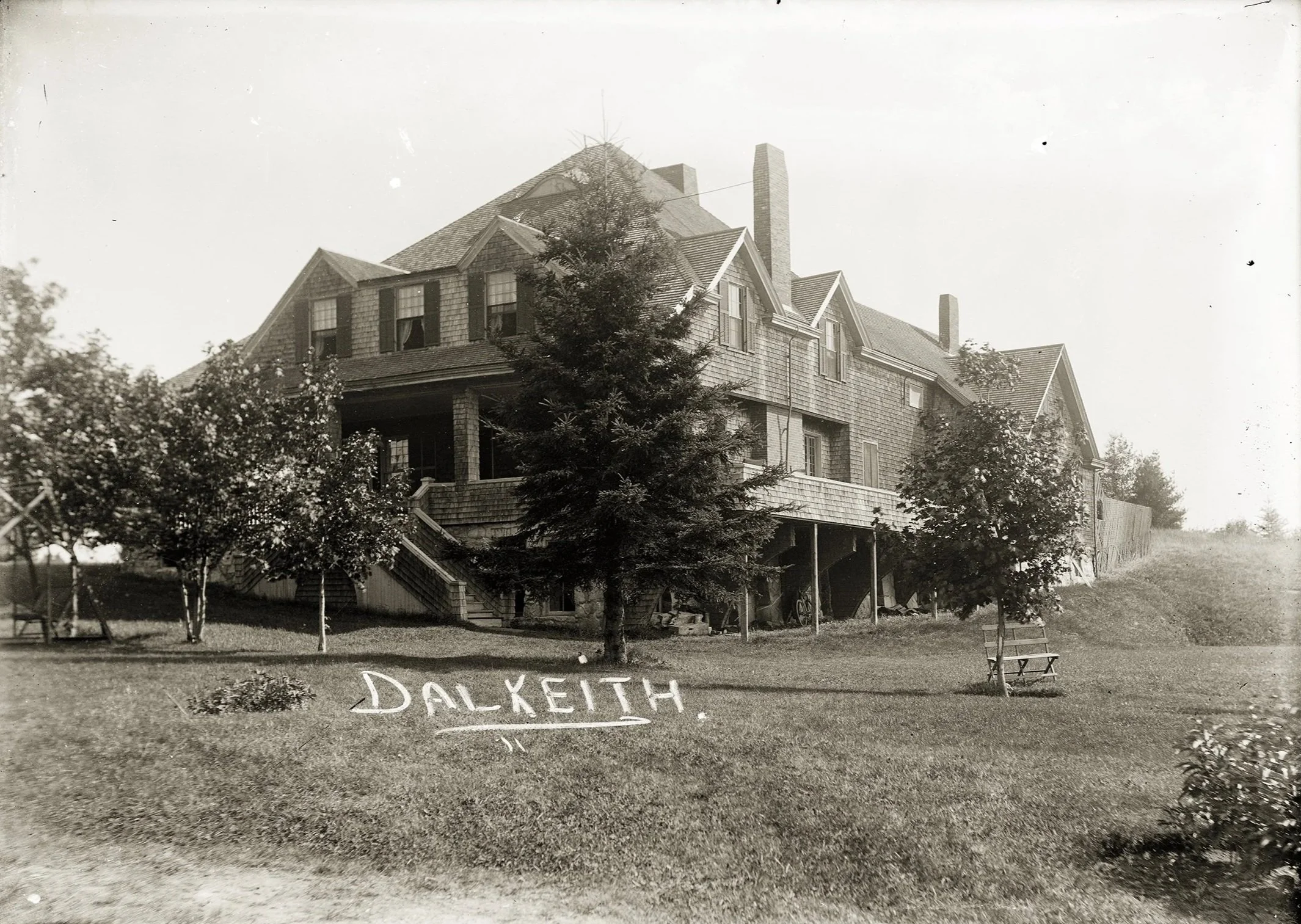 A black and white photo of a large, old house with steep roofs, multiple chimneys, and a porch. In front of the house, there are several trees, a patch of grass and some benches and chairs.