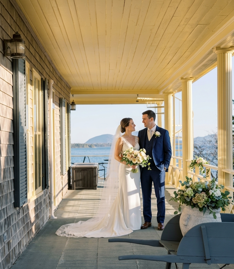 Bride and groom standing on a porch overlooking the water, smiling at each other, with wedding flowers and decor around.