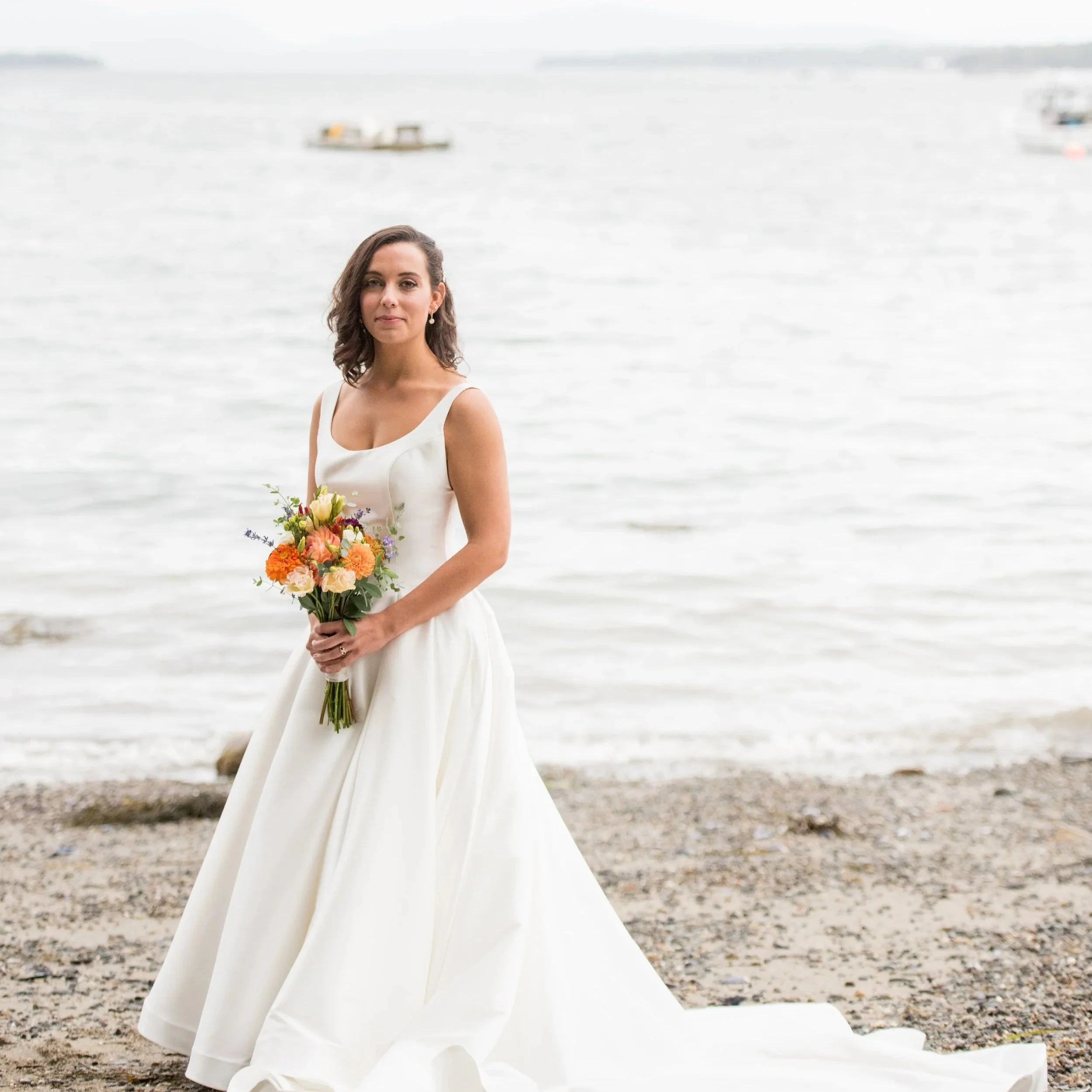 A woman in a white dress holding a bouquet of flowers on a beach with water and boats in the background.