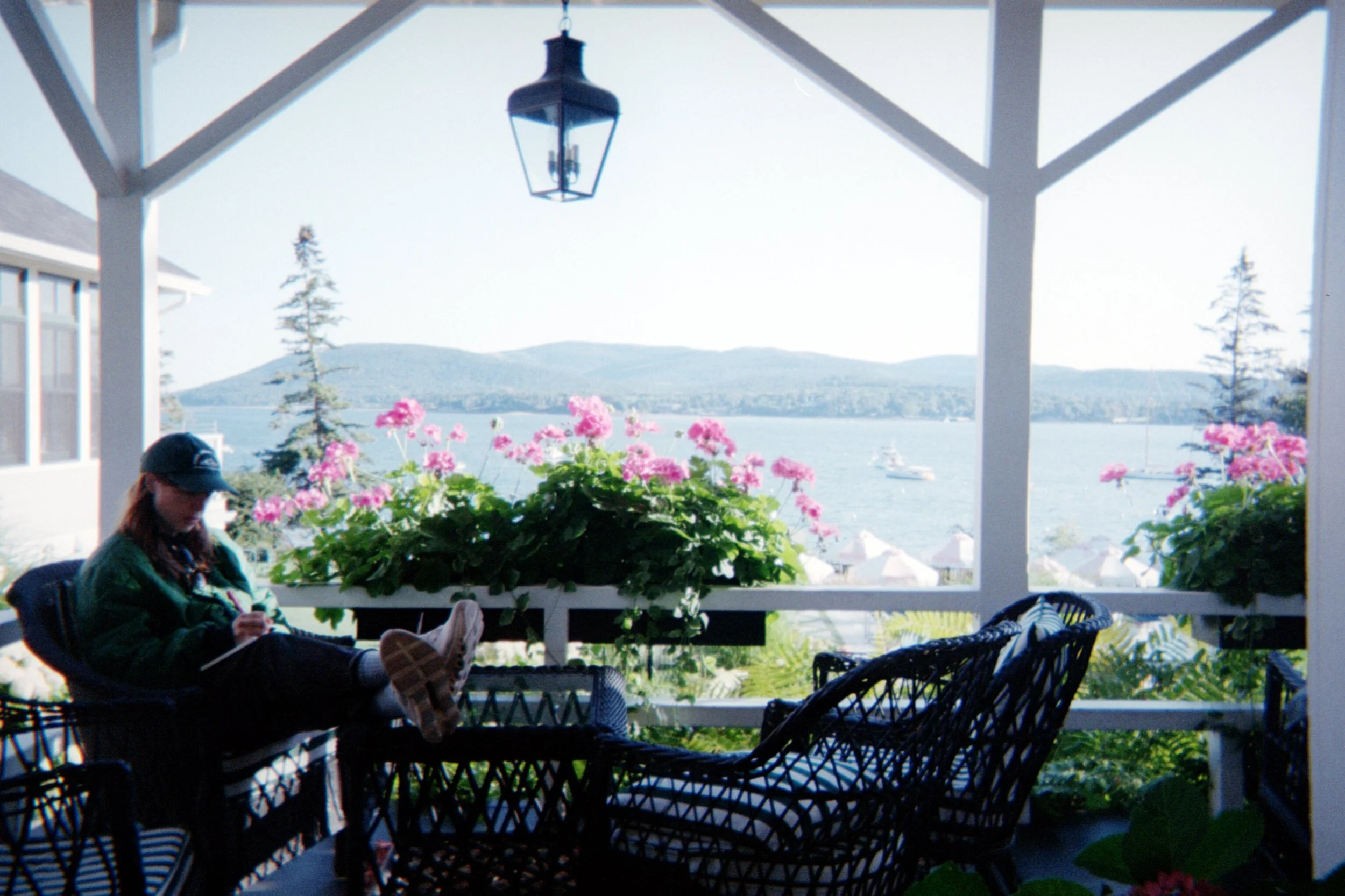 A covered porch overlooking a lake with mountains in the distance. There are pink flowers in the foreground, a hanging lantern above, and a person sitting on a chair wearing a cap and jacket.