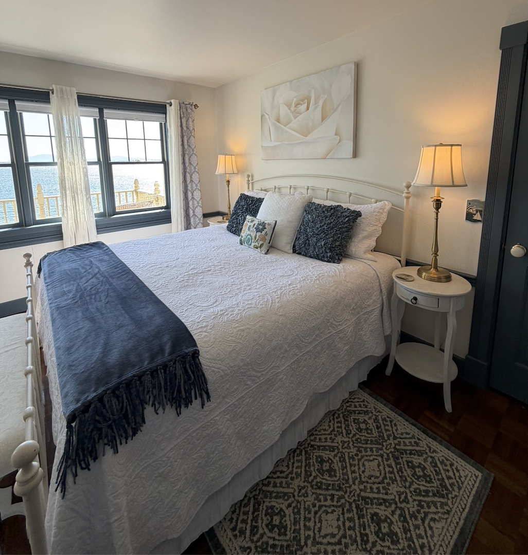 A cozy bedroom with a large bed covered in a white quilt and decorative pillows. There is a navy blue throw at the foot of the bed. On each side of the bed are matching bedside tables with lamps. The room has a large window with white and patterned curtains, offering a view of a body of water and a railing outside. A large floral painting hangs above the bed, and a patterned rug is partially visible on the dark wooden floor.