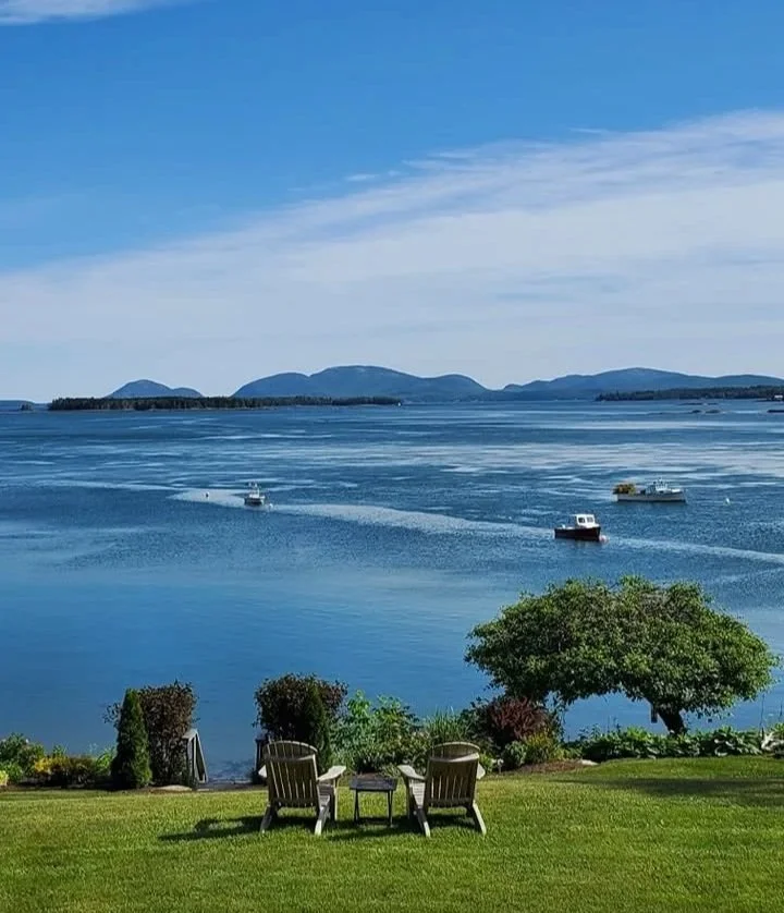 A scenic view of Frenchman's Bay, Bar Harbor, and Acadia National park with boats and distant mountains in the background, seen from a lawn with chairs and landscaping at the Acadia Bay Inn Bed & Breakfast.