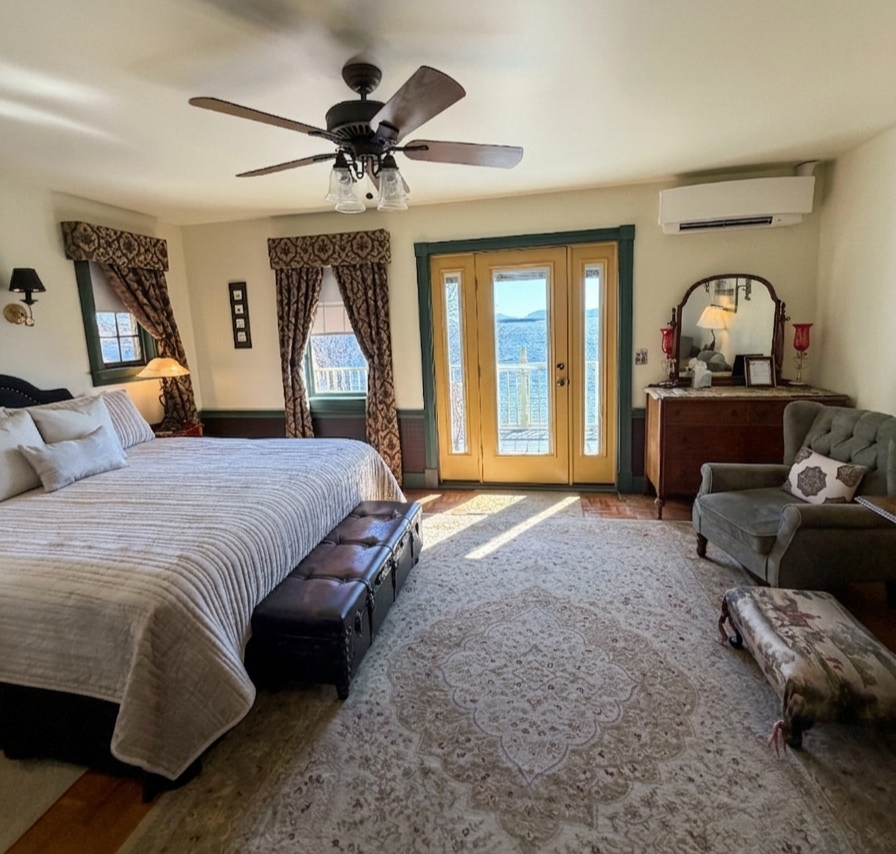 A bedroom with a large bed, wooden furniture, windows with drapes, a ceiling fan, and a door leading outside showing a view of water and sky.