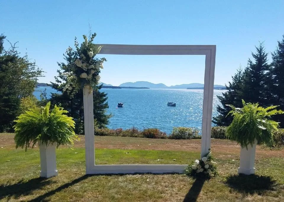 A decorative white frame set up outdoors with two potted ferns and floral arrangements, overlooking a body of water with boats, trees, and mountains in the background on a sunny day.