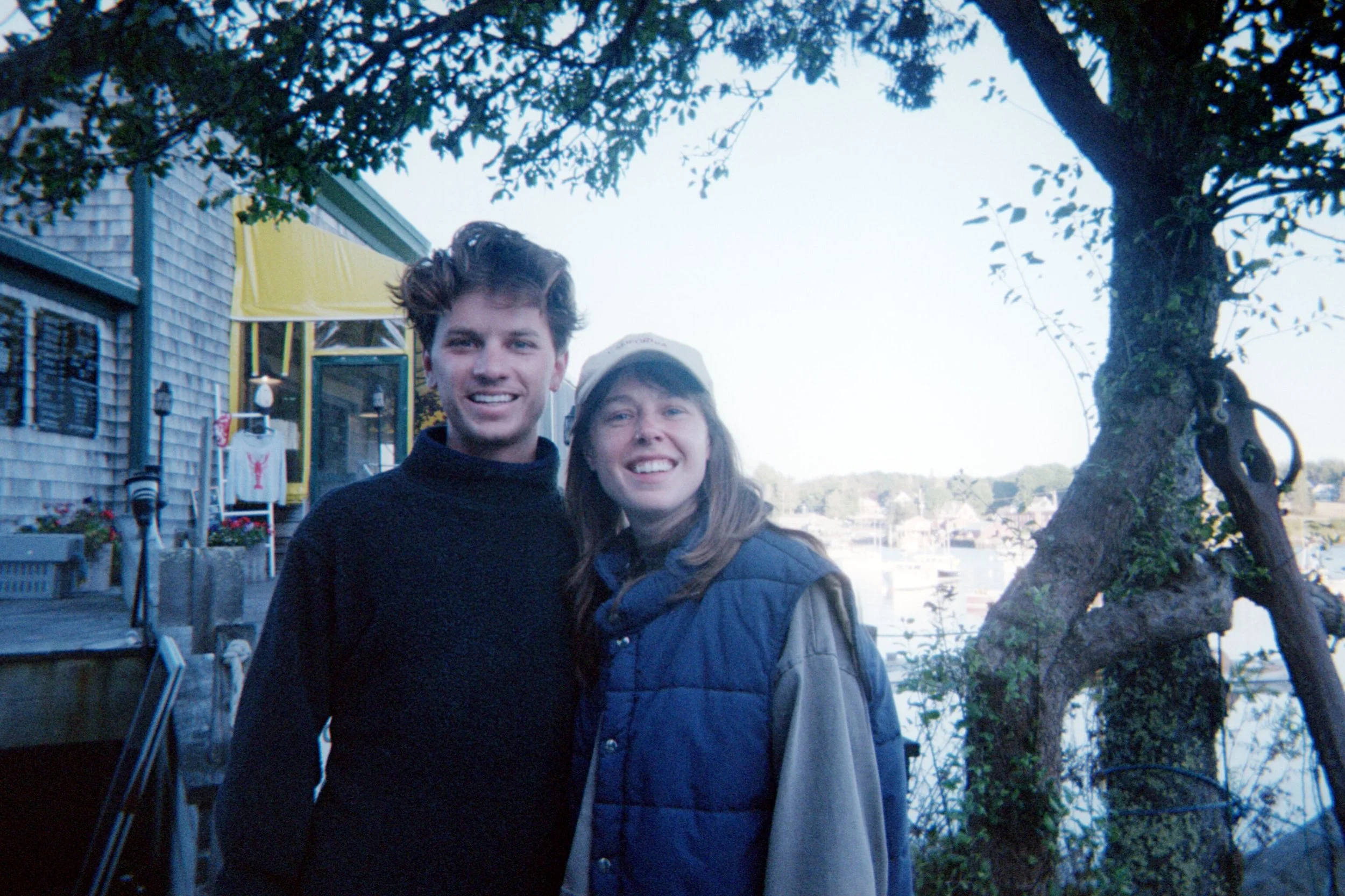 A young man and woman smiling outdoors near a tree, with a house and a canal in the background.