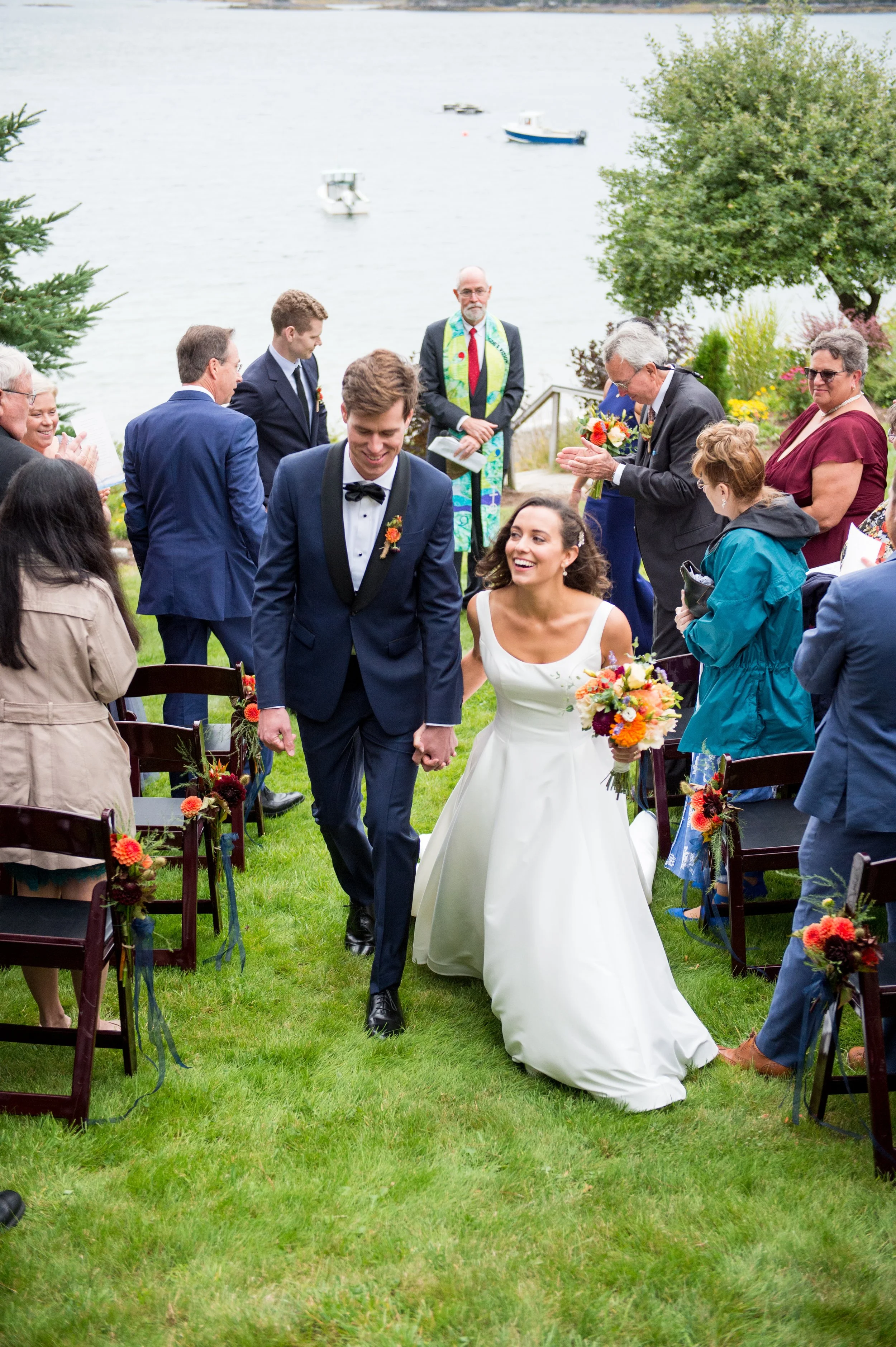 Bride and groom walking hand in hand down the aisle at an outdoor wedding ceremony by a body of water, surrounded by guests and floral decorations.
