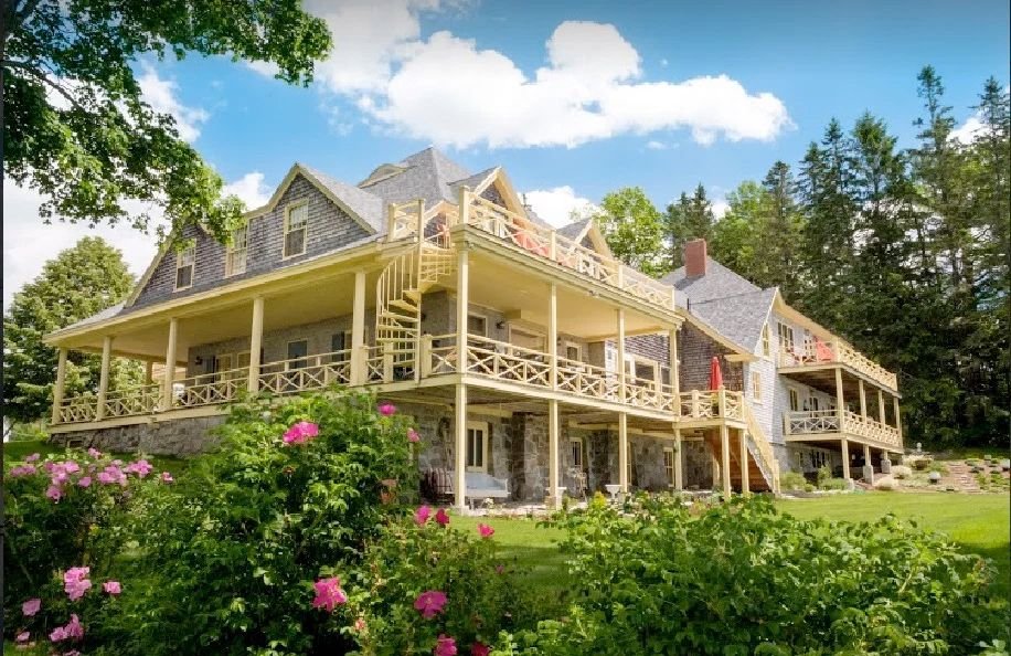 A large multi-story shingle-style inn with stone and cedar shingle exterior surrounded by lush green trees and pink flowers under a partly cloudy sky.