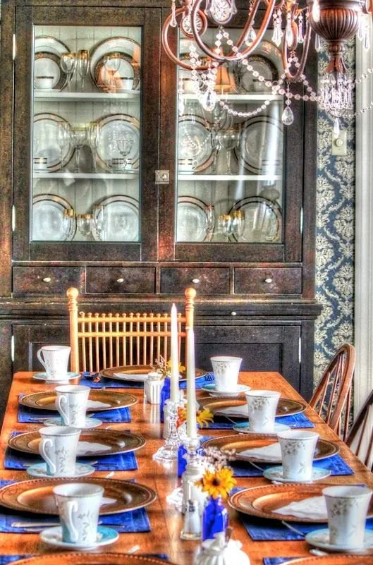 A wooden dining table set for eight, with copper-colored plates, white teacups, blue napkins, and silverware. In the center, there are tall candlesticks with white candles, small vases with yellow flowers, and blue ribbon accents. Behind the table is a black china cabinet displaying plates and bowls. A crystal chandelier hangs overhead, illuminating the setting.
