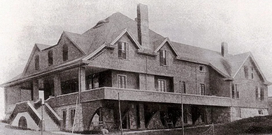Black and white photo of a large, historic house with multiple stories, steep gable roofs, numerous windows, and a wraparound porch supported by columns.