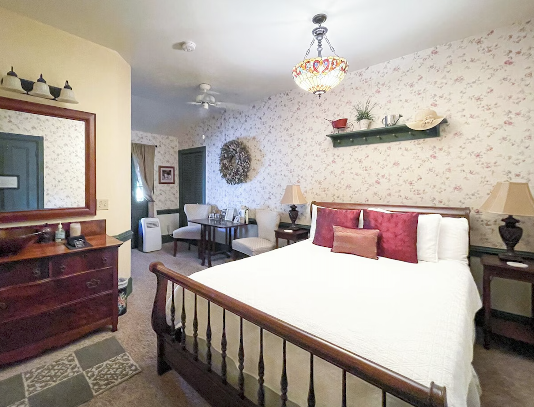 Hotel room with a bed, nightstands, lamps, wall decor, and vintage furniture, floral wallpaper, chandelier, ceiling fans, and window with curtains.