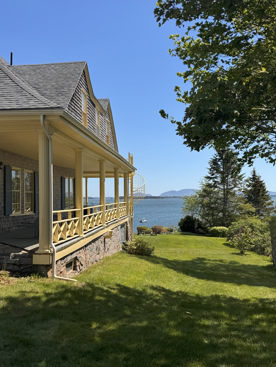 A waterfront inn with a second-story deck overlooking Acadia National Park, surrounded by lush green grass and trees, under a clear blue sky.
