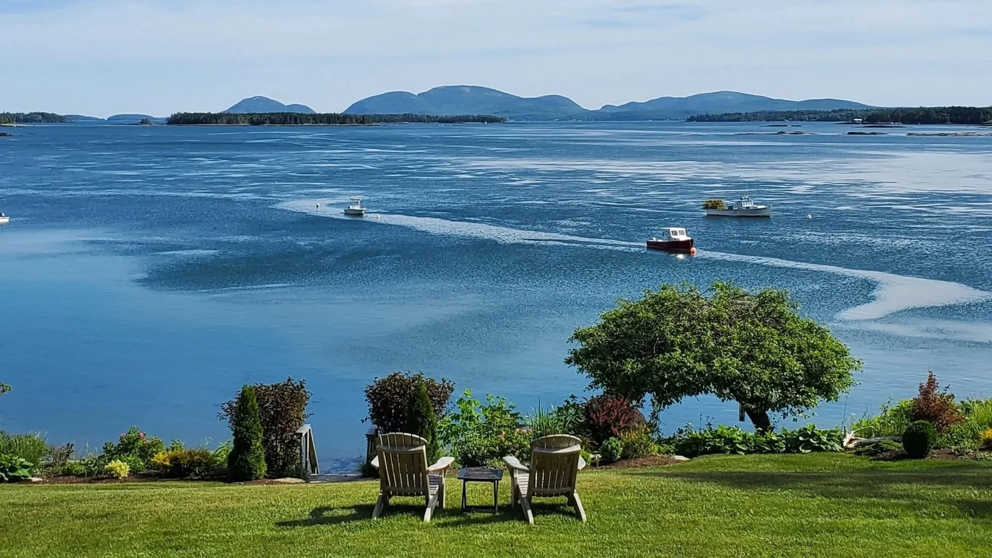 A scenic view of Frenchman's Bay, Bar Harbor, and Acadia National park with boats and distant mountains in the background, seen from a lawn with chairs and landscaping.