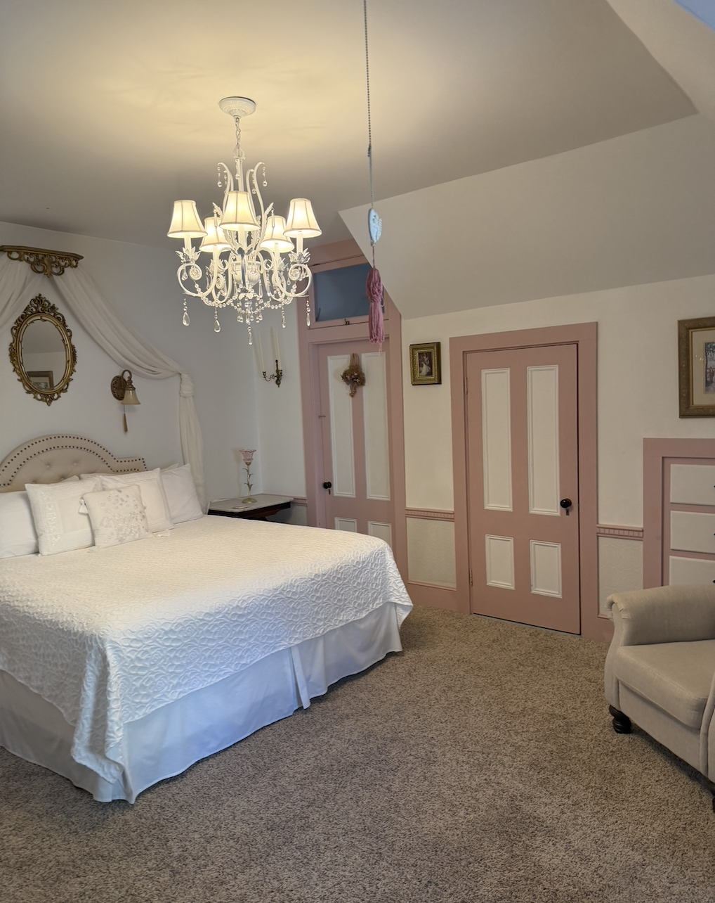 A vintage bedroom with a large bed covered in white bedding, a chandelier hanging from the ceiling, a bedside table with a pink lamp, a beige armchair, pink and cream doors, and framed artwork on the walls.