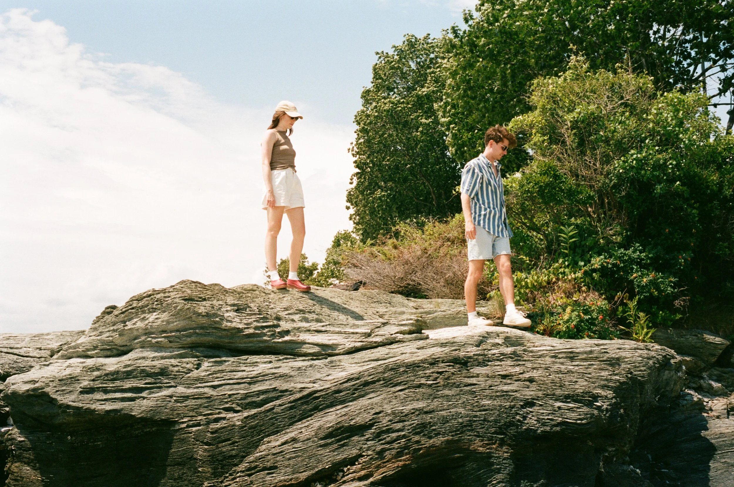 Two people are walking on a large, flat rock formation outdoors near some bushes and trees, under a partly cloudy sky.