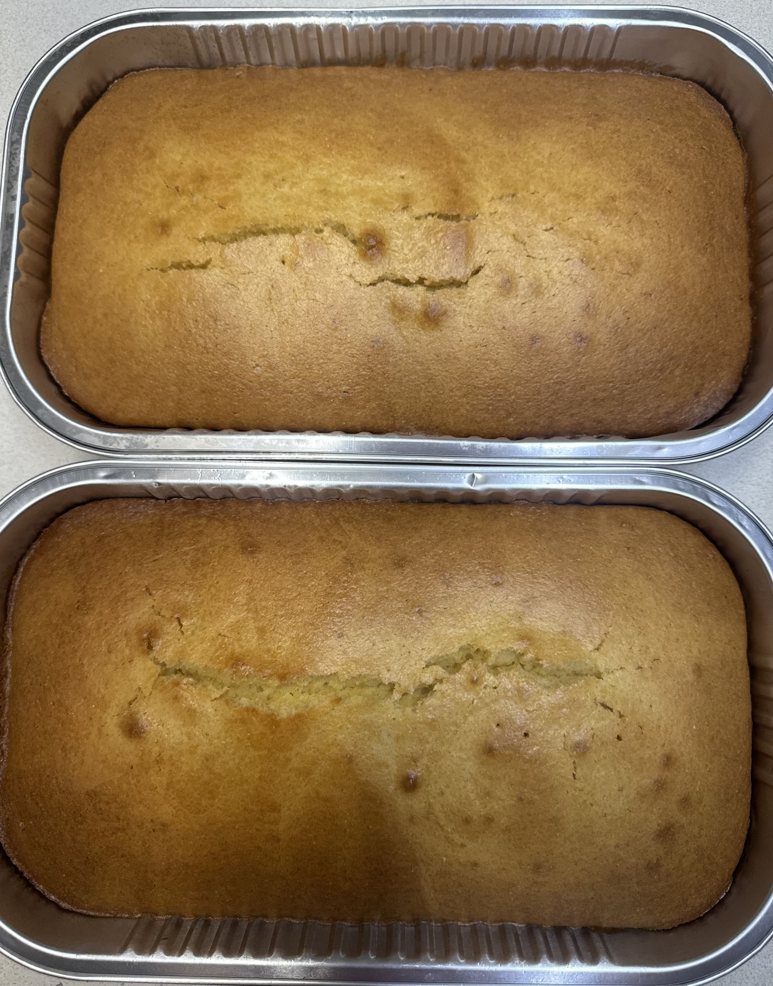 A close-up of two large rectangular baked cakes in metal pans, with one cake showing a slightly cracked surface.