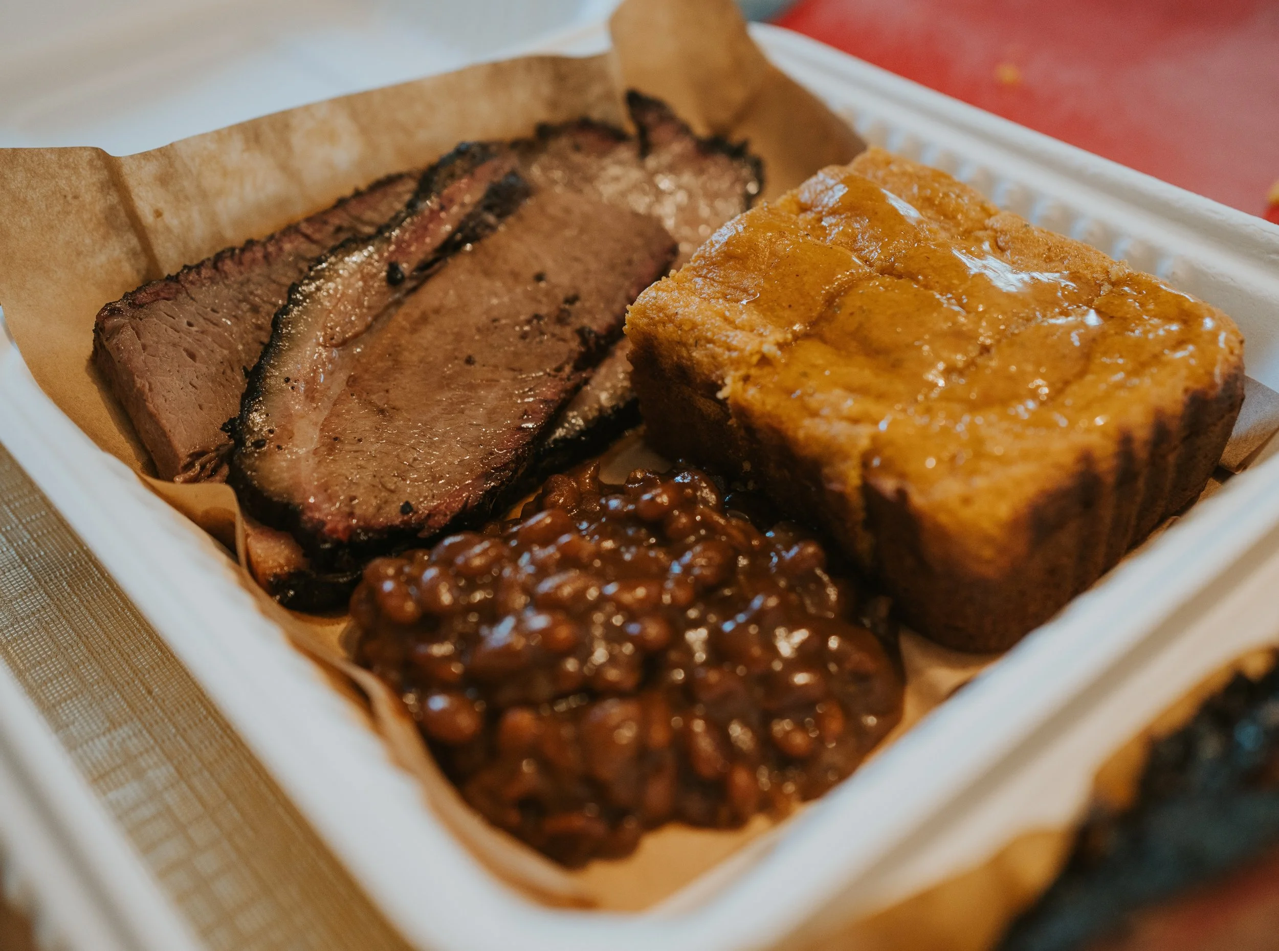 Slices of cooked brisket, baked bean casserole, and cornbread in a takeout container.