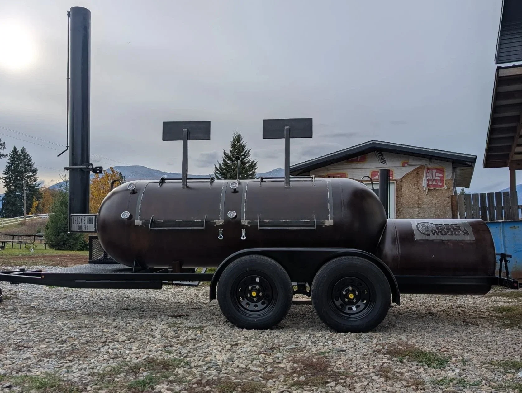 A black barbecue smoker on a trailer, located outdoors on a gravel surface with trees, mountains, and a cloudy sky in the background.