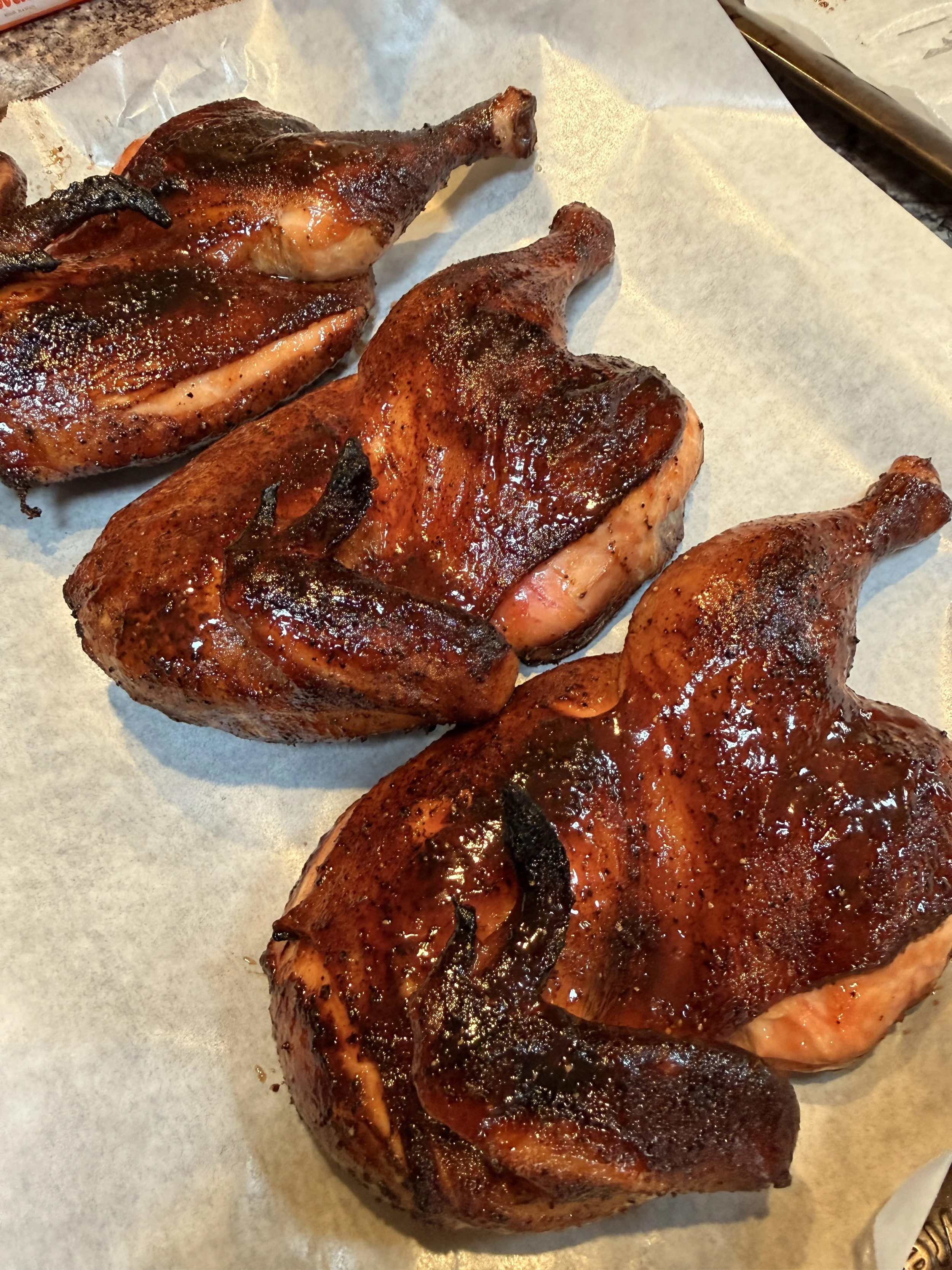 Three cooked chicken leg quarters with a dark, crispy marinade, placed on a sheet of parchment paper on a countertop.