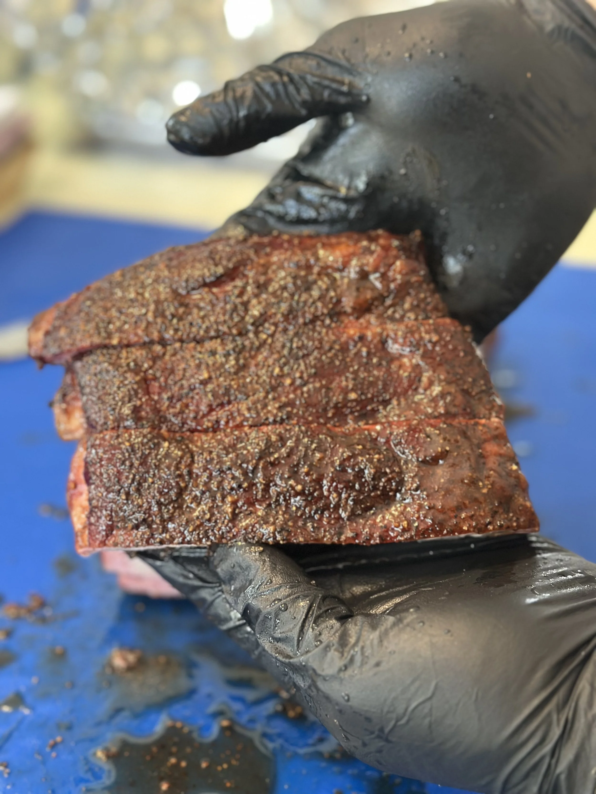 A close-up of two pieces of raw seasoned meat held by a person wearing black gloves, with a blue cutting board and some scattered seasoning in the background.