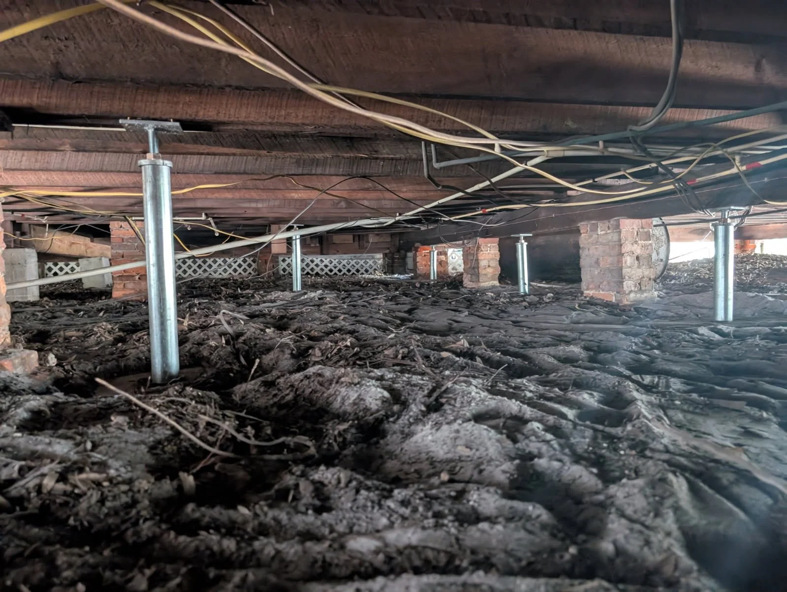 Underneath a raised house, showing a dirt floor, supporting poles, and electrical wiring running along the wooden beams.