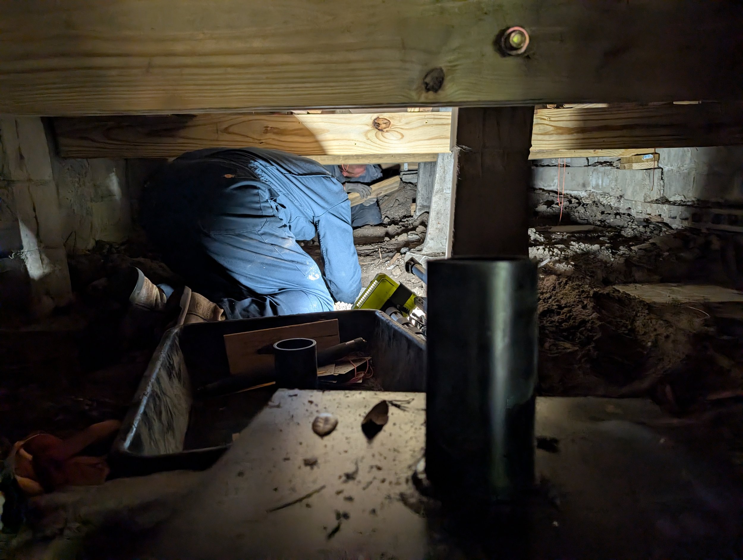 Person working on a construction site, crouched down and wearing a blue jacket, surrounded by wooden beams and soil. fixing floor joists and crawlspace floors